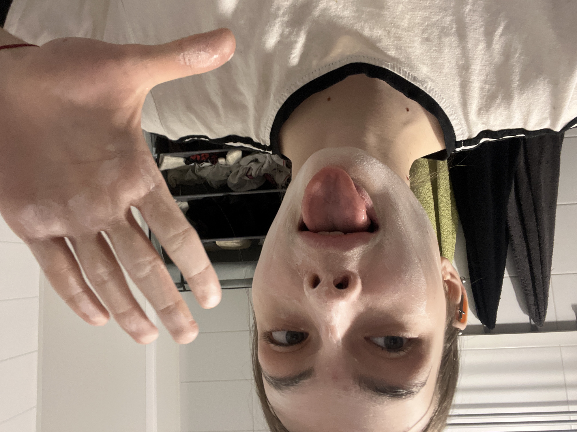 This close-up, horizontally flipped and upside-down image, depicts a person, likely young, engaged in a self-care routine, possibly applying a face mask or lotion. The individual's face and extended right hand are covered in a white, creamy substance. The person is playfully sticking their tongue out, with eyes slightly looking upward.
They are wearing a white t-shirt with a black trim around the collar. A thin red string or bracelet is visible on their right wrist, and their left ear features multiple piercings. The background suggests an indoor bathroom setting, with white tiled walls, a dark towel hanging, and a shelf with folded items (possibly clothes or towels) partially visible. The lighting is artificial, characteristic of indoor evening or night conditions. No text is visible in the image. The scene takes place in Saint Petersburg, Russia.