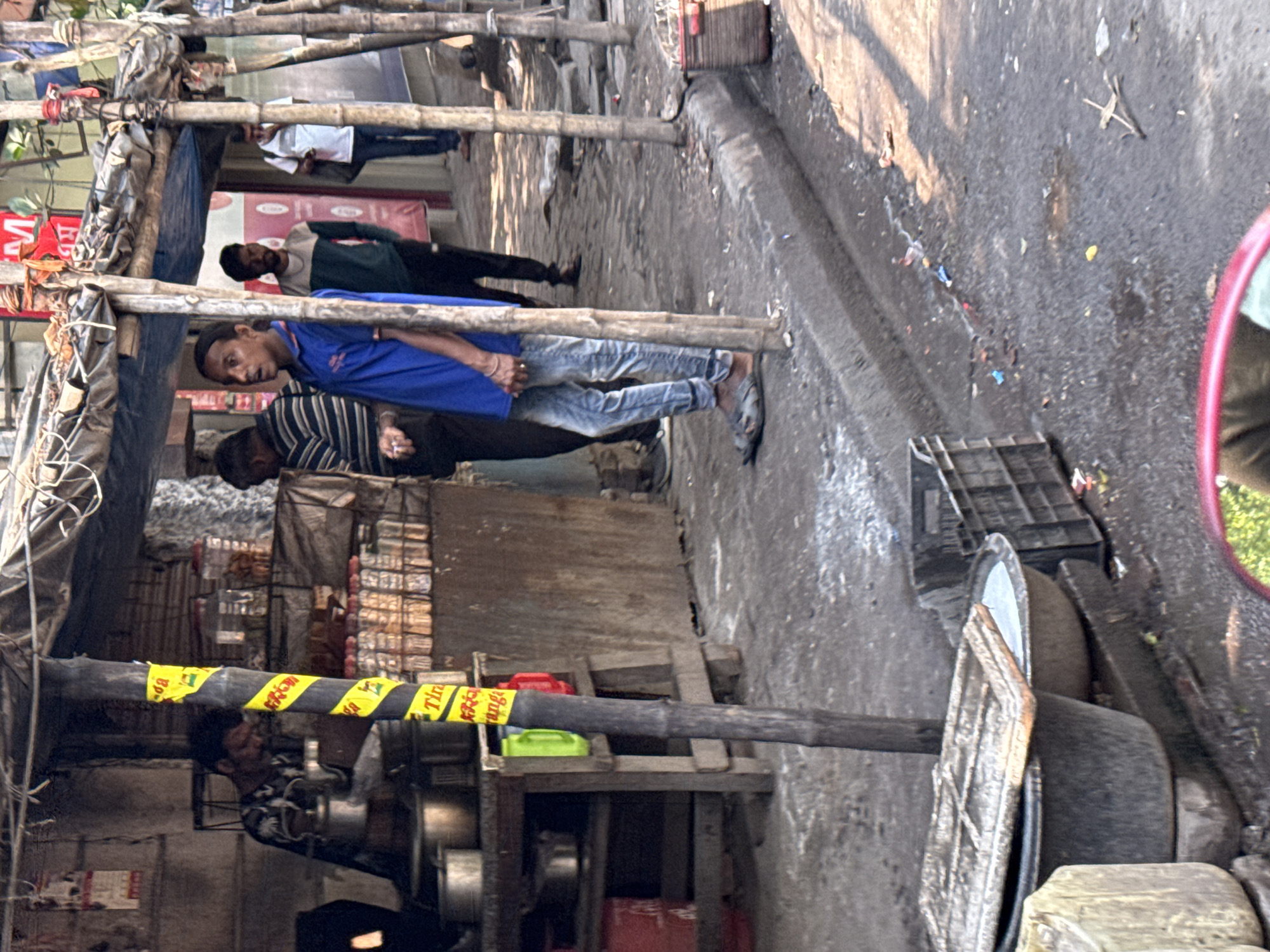 This image depicts an active street scene in Kolkata, India, characterized by informal commerce and pedestrian movement under bright daylight.
In the foreground, a dark grey basin with a partial wooden lid sits on the rough concrete ground, containing water. Adjacent to it are a smaller basin, also containing water, and a dark plastic crate. The ground exhibits dirt, scattered refuse, and damp patches. A red-framed object, likely a vehicle mirror, encroaches from the bottom right edge.
The midground features a makeshift food or tea stall on the left, sheltered by a blue tarpaulin canopy supported by wooden and bamboo poles. A central bamboo pole is wrapped with yellow and black striped tape, displaying partial text including "Tira Orange". A man with dark hair and a beard, wearing a patterned shirt, operates the stall, leaning over a stove with multiple metal pots and kettles. Shelves behind him hold various packaged snacks and bottled items. To the left of the central pole, another man in a striped shirt stands, facing the stall.
To the right of the stall, a young man or boy, wearing a blue polo shirt, light blue jeans, and dark sandals, stands on the curb, looking intently towards the right with an open mouth. Further back on the street, another man in a green and white striped shirt stands. Beyond him, a man in a white t-shirt with a dark bag walks away from the viewer. The street surface is rough, showing dirt and debris.
In the background, buildings with visible windows and faded signage, including red text, line the street. Sparse foliage is visible above the structures. The scene is cast with strong shadows, indicating direct sunlight.