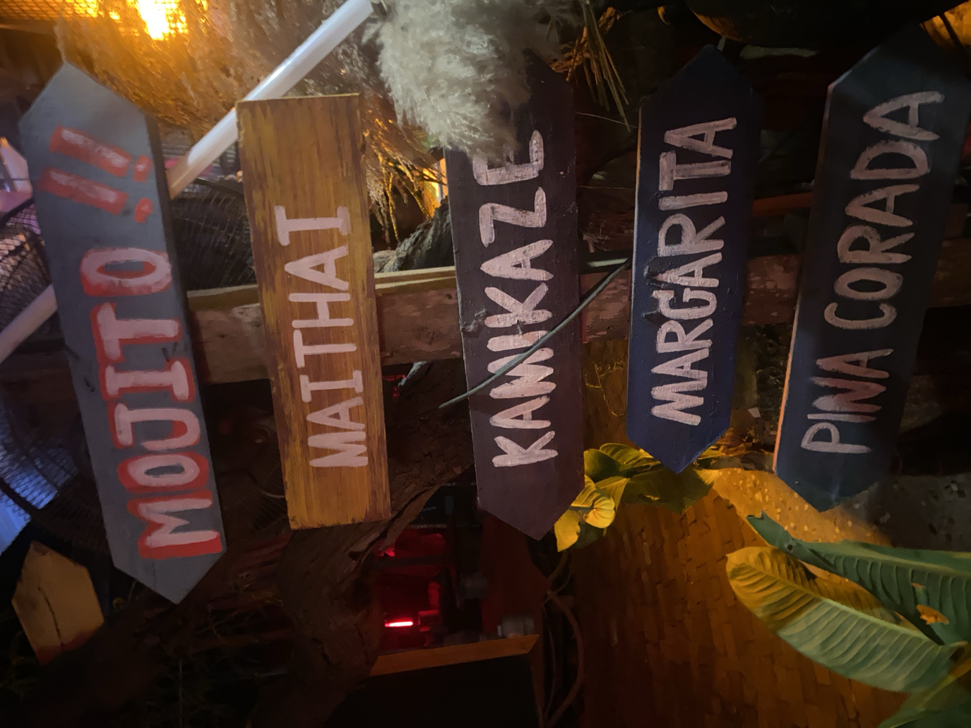 Five signs for cocktails are arranged in a row. The signs are made of wood and painted with the names of the cocktails in white lettering: "Mojito," "Maithai," "Kamikaze," "Margarita," and "Pina Corada." The signs are situated among greenery and appear to be part of a bar or restaurant setting. The location is Baan Don Sai, Thailand.