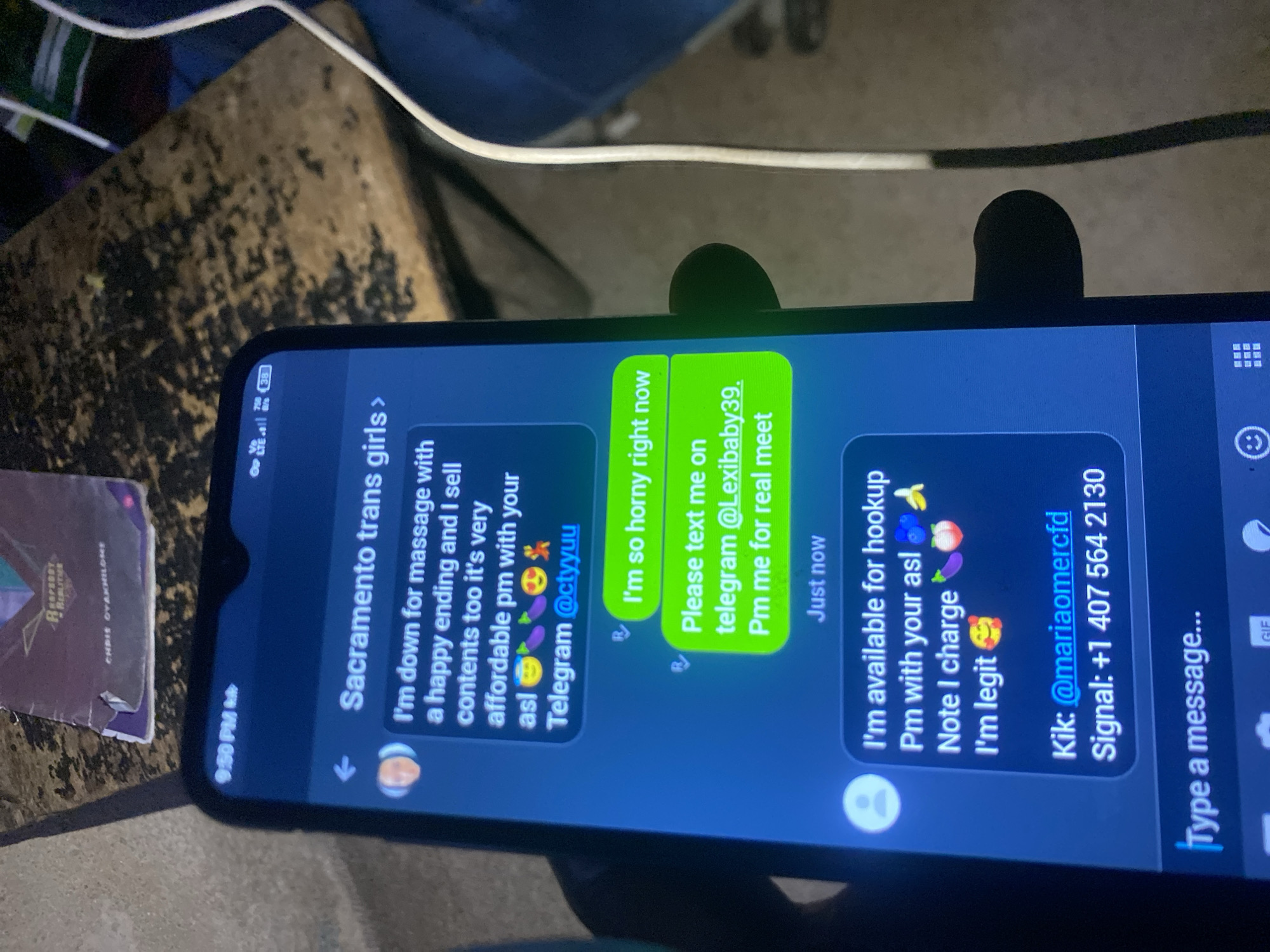 The image shows a close-up of a smartphone screen displaying a Telegram chat with a group named "Sacramento trans girls." The chat messages suggest the individuals are offering or seeking sexual encounters, referencing massage with a happy ending, selling content, and hookups. Contact information, including usernames for Telegram and Kik, along with a phone number for Signal, are visible in the messages. The time displayed on the phone is 9:50 PM. The image appears to be taken indoors, with a glimpse of a table surface and a partially visible book cover in the background. There is no explicit information in the image to confirm that the location is in the city of Bukuru, Nigeria.