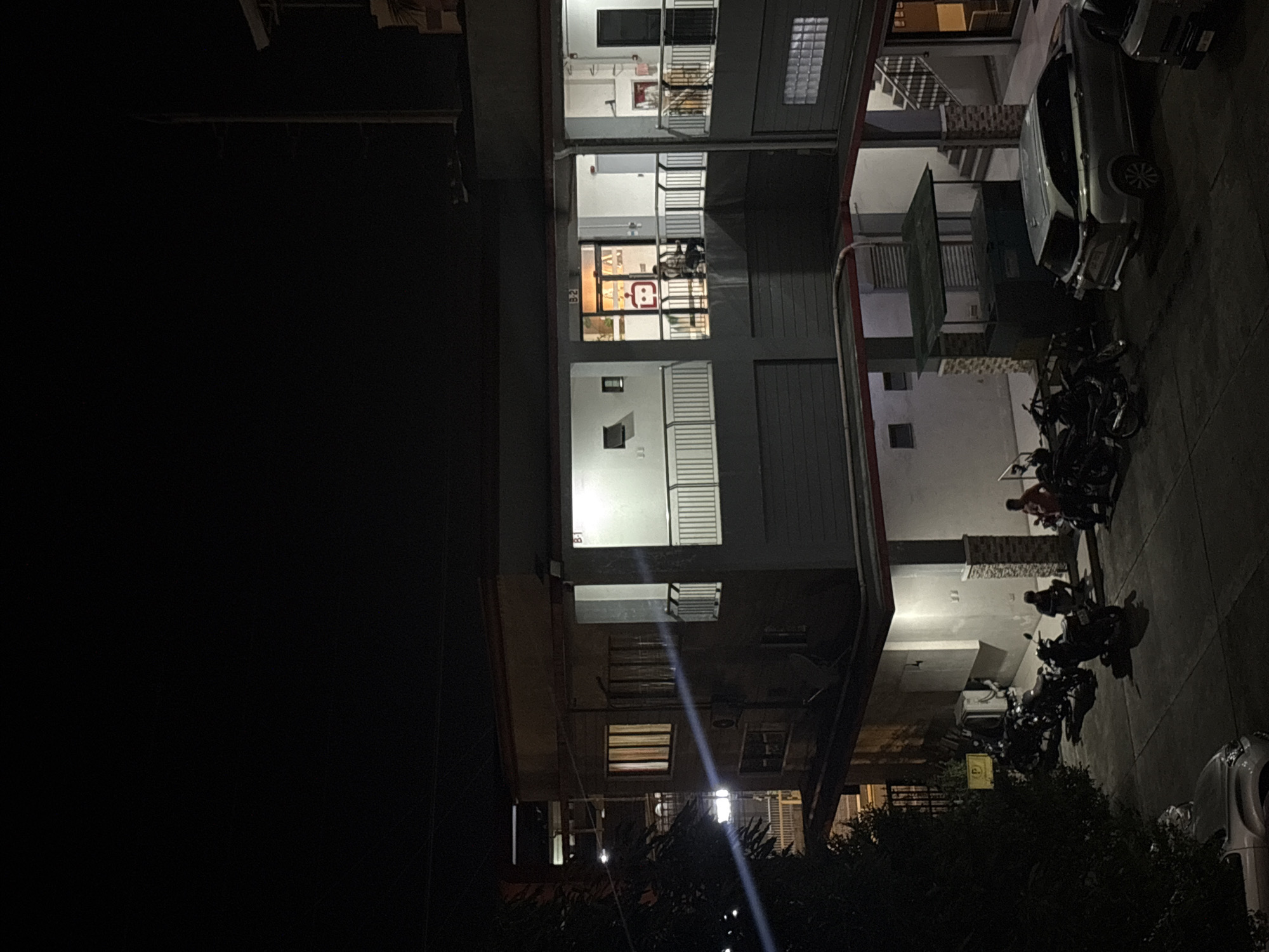 The image captures a nighttime scene in Davao City, Philippines, likely a residential or commercial complex. The primary focus is a multi-story building with illuminated windows and balconies. In the foreground, several motorcycles are parked on a paved area, with two individuals seated near them. A silver SUV is also visible, parked to the right of the motorcycles.
The building itself appears to have a modern design with a grey facade and white balconies. Some windows show warm interior lighting, suggesting occupants are present. A small guard house or kiosk is situated near the parked vehicles. The overall environment is dark, illuminated by artificial lights from the building and streetlights. The presence of motorcycles and an SUV suggests a functional area for residents or patrons. The image's perspective is from an elevated viewpoint, possibly a window or another building across the street.