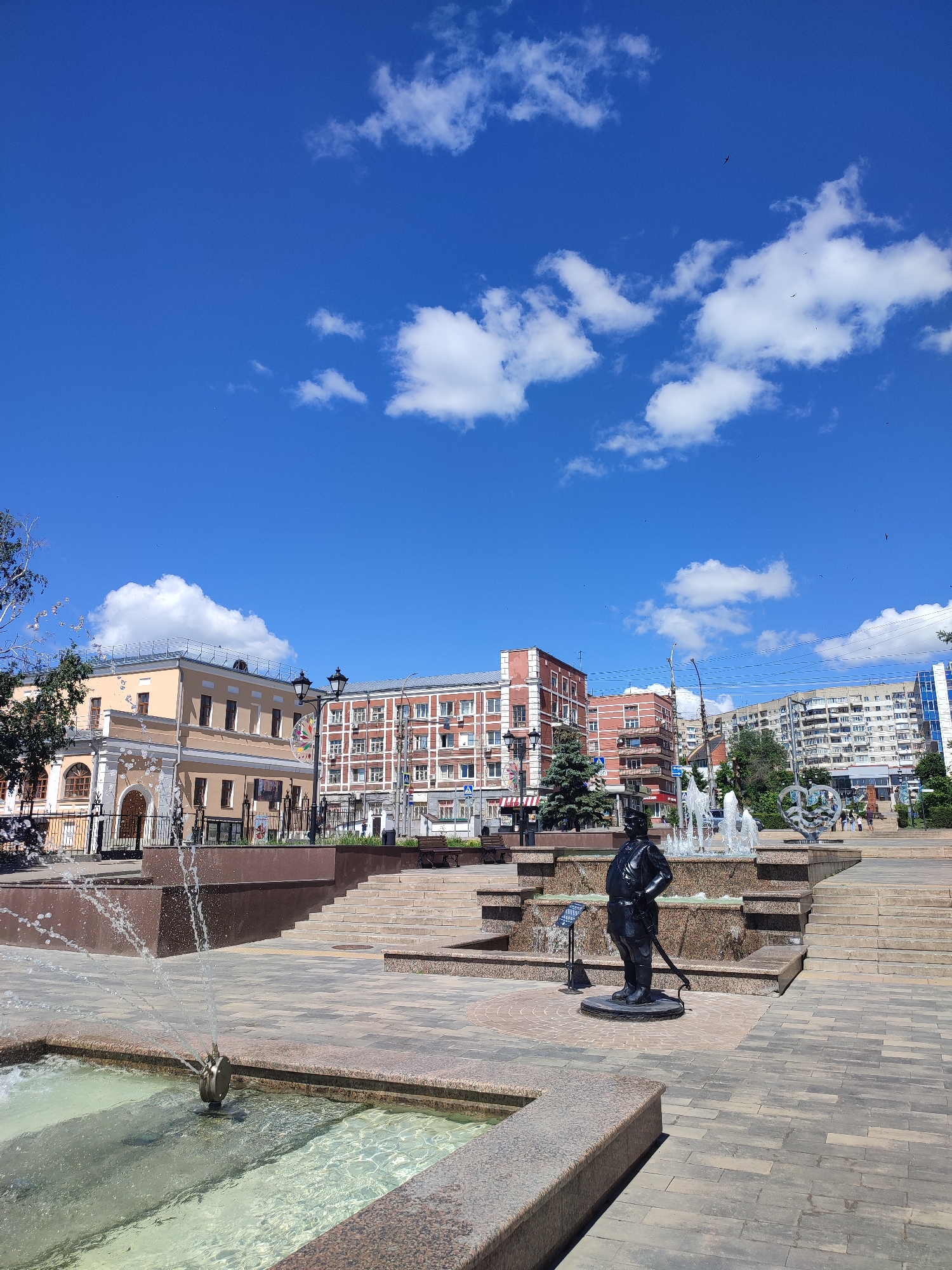 This is a vertical, eye-level, full shot of a public square in Saratov, Russia, on a bright, sunny day. The sky is a vibrant blue with scattered white cumulus clouds, and a few dark specks that may be birds or distant aircraft are visible. In the foreground, a multi-tiered fountain is active, with water spraying upwards and cascading down. A dark statue of a man in a uniform, possibly a military figure, stands on a pedestal in front of the fountain, with a sword by his side. The square is paved with light-colored interlocking bricks and granite.
Behind the fountain, a wide staircase leads up to a higher level of the square. In the background, several buildings are visible. To the left, a light yellow, two-story building with arched windows and a fence in front is prominent. To its right, a red-brick building with many windows stands tall. Further back, a modern, multi-story apartment building dominates the skyline. Trees are interspersed throughout the scene, with a dark green evergreen tree standing near the statue and a leafy tree with falling water droplets visible on the far left.
The time of day appears to be midday, given the bright sunlight casting sharp shadows. The overall mood is cheerful and lively, suggesting a pleasant day for outdoor activities. The presence of benches and the well-maintained public space indicate it's a popular gathering spot. No visible text is discernible in the image.