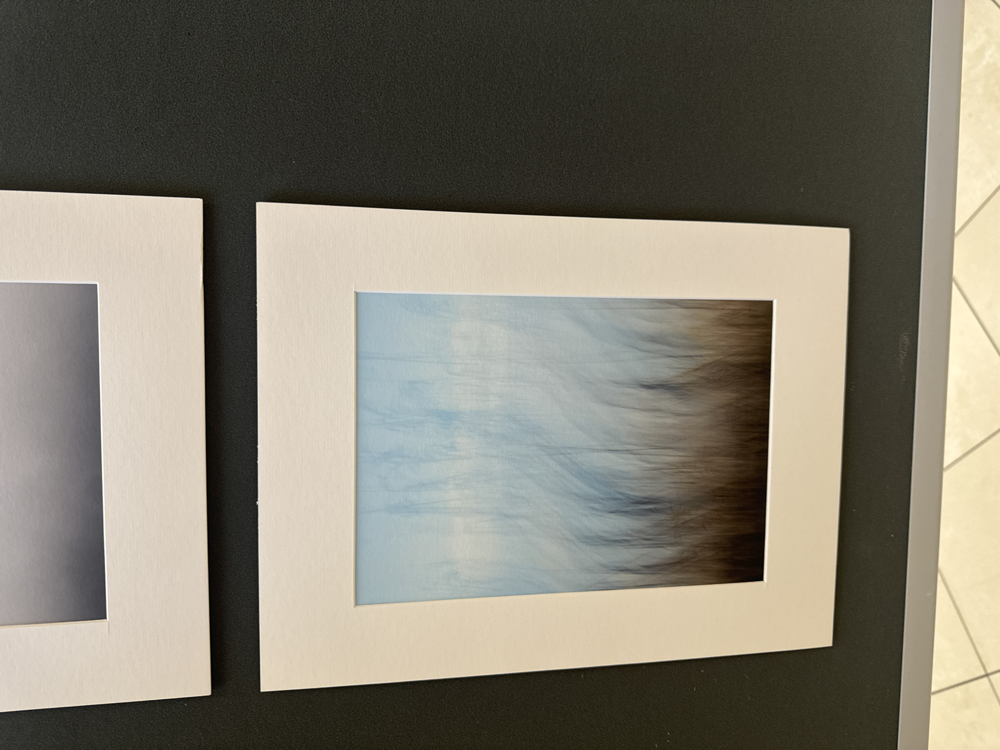 This image shows two matted photographs displayed on a dark gray wall. The top photograph is partially visible and appears to be a landscape with a gradient of grays. The bottom photograph is fully visible and shows an abstract impression of tall reeds or grasses against a pale blue sky. The bottom photograph is matted in off-white. The wall has a textured surface and is mounted on a gray baseboard. The floor below is tiled with a light beige and white pattern. The image is taken at eye level, looking slightly upwards. This scene appears to be an indoor exhibition space, possibly in a gallery or public building. There are no people or animals visible. It is not possible to determine the location in the city of Kilkenny, Ireland from the provided image.