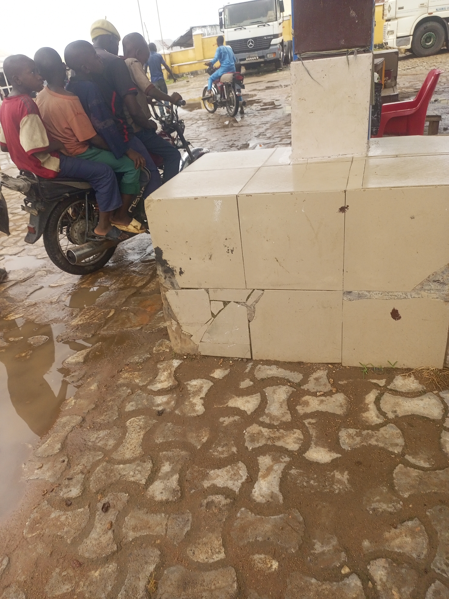 In the city of Serti, Nigeria, six young boys are precariously perched on a single motorcycle. The rider, wearing a yellow cap, is in the front, followed by five boys crammed together. The motorcycle is parked on a wet, cobblestone-like surface, indicating recent rainfall. The boys, dressed in casual clothing, appear to be waiting or have just disembarked. In the background, a white truck and a red plastic chair are visible, suggesting a commercial or roadside setting. The overall scene depicts a common mode of transportation and the everyday life of the community in Serti.