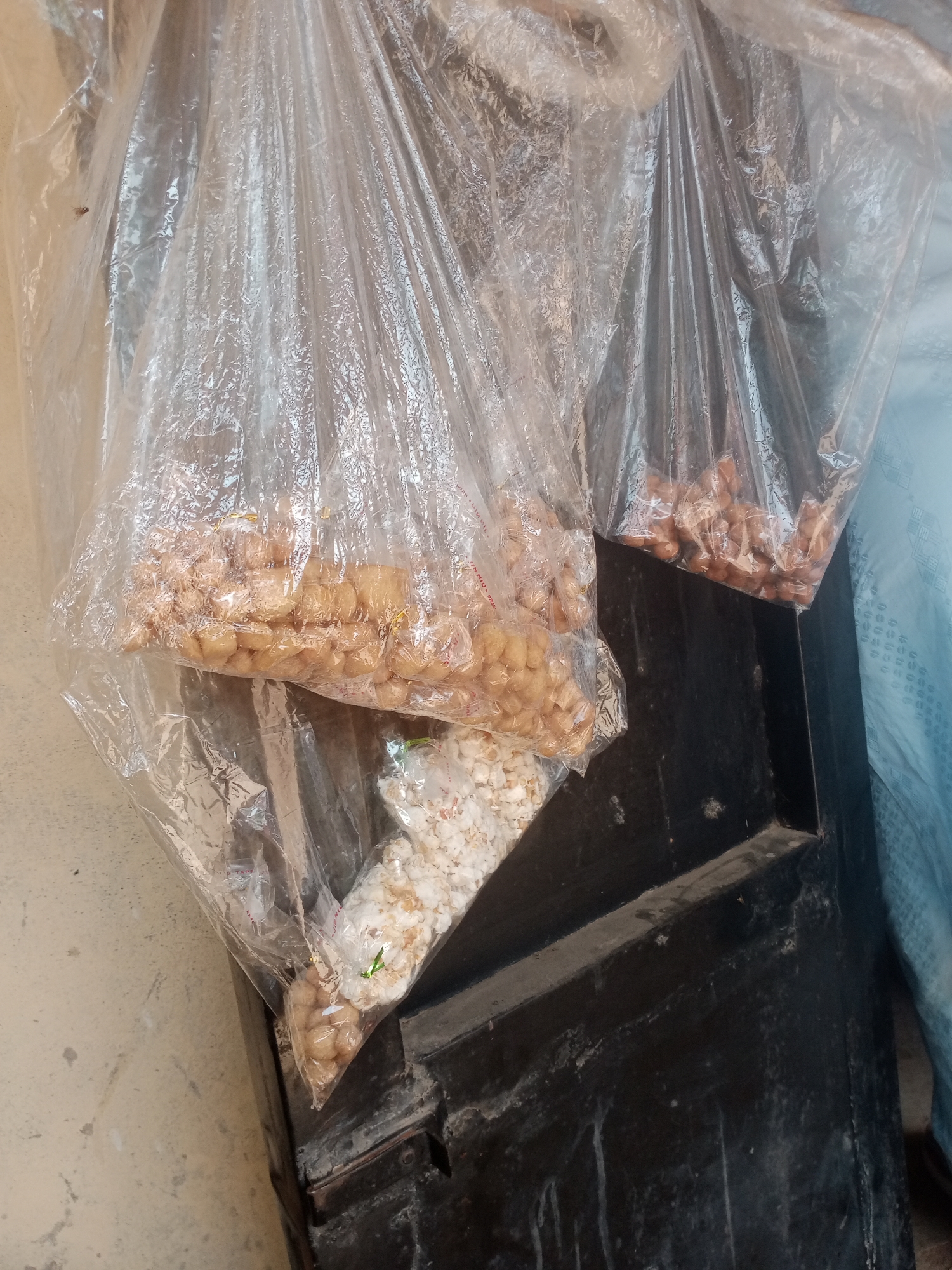 The image depicts a close-up view of several clear plastic bags filled with various snacks, seemingly displayed for sale. Two larger bags, on the left and right sides of the frame, are filled with small, light-brown, cube-shaped snacks. A third, smaller bag, positioned below the left bag, contains white, puffy snacks that resemble popcorn. All the bags are hanging and appear to be somewhat transparent, allowing the contents to be clearly visible. The bags are placed against a dark, likely wooden, surface, possibly a door or a stall. To the left of the bags, a textured, light-colored surface, perhaps a wall or concrete, is visible. The lighting suggests an outdoor or well-lit indoor environment. The overall scene conveys a market or street vending setting. The text visible on one of the bags, "JINNIU", suggests a possible brand or origin. The image was taken in Jalingo, Nigeria.