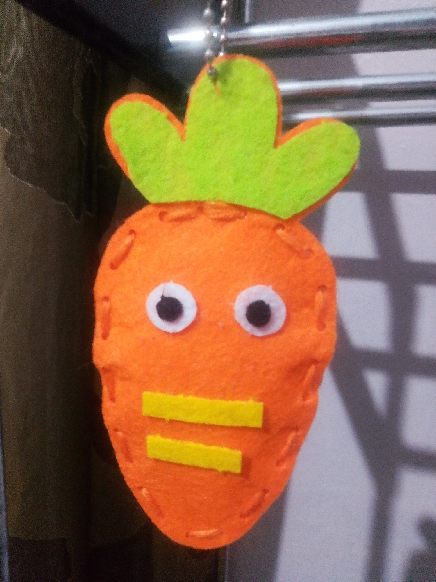 A close-up, eye-level shot shows a handmade felt carrot ornament hanging from a metal bar. The ornament is orange, carrot-shaped, with green felt leaves at the top. Two white circles with black centers represent eyes, and two horizontal yellow felt rectangles form a mouth. The carrot has visible orange stitching around its edges. The background is blurred, with a dark, textured fabric on the left and a light gray surface with a grid-like shadow pattern on the right. The ornament is suspended by a silver chain. No specific location in Tekeli, Kazakhstan is discernible from the image.