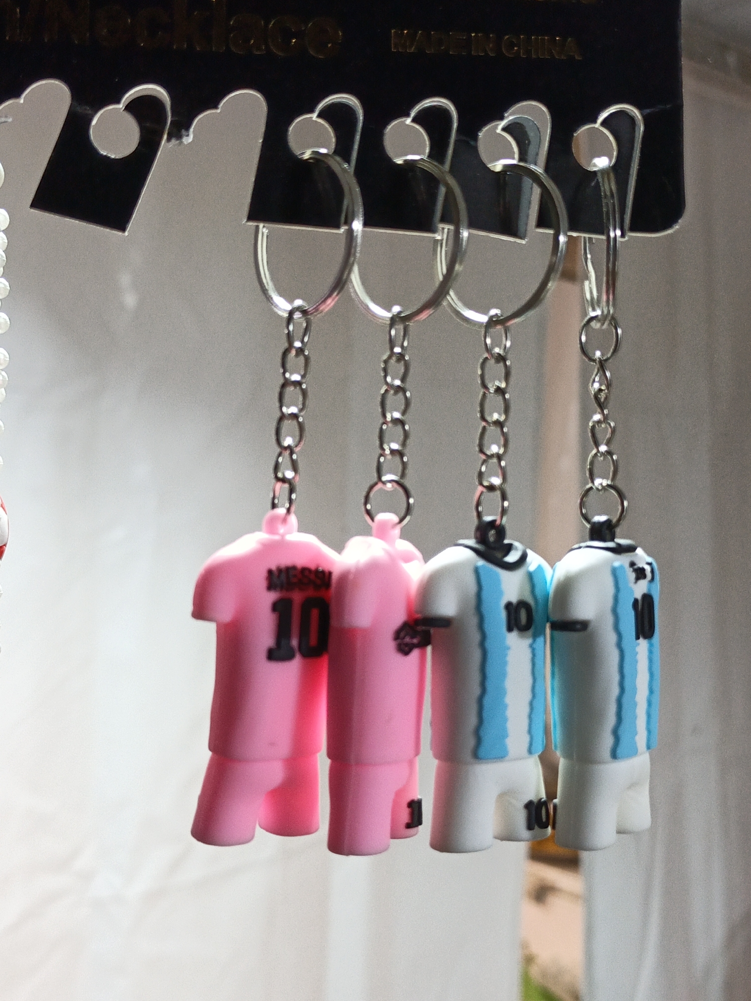 This media file shows a close-up of four keychain fobs, each designed to resemble miniature soccer jerseys. The jerseys are attached to metal keychains with small circular rings and clasps, which are hanging from a black backing card. The backing card has text printed in gold, with "Necklace" visible on the left and "MADE IN CHINA" on the right.
The keychains themselves are of two types. The two on the left are bright pink and feature the name "MESSI" and the number "10" in black lettering on the back. The two on the right are white with light blue vertical stripes, and also have the number "10" printed in black. All four jerseys have black collars. The keychains are hanging in a row, suggesting they are part of a product display.
The background is softly blurred and appears to be a plain, light-colored surface, possibly a wall or fabric. There is no indication of the time of day, weather, or a specific geographical location. The lighting is even, and the focus is entirely on the keychains, creating a sense of it being a product shot. The scene is static, with no activity taking place other than the display of the objects.