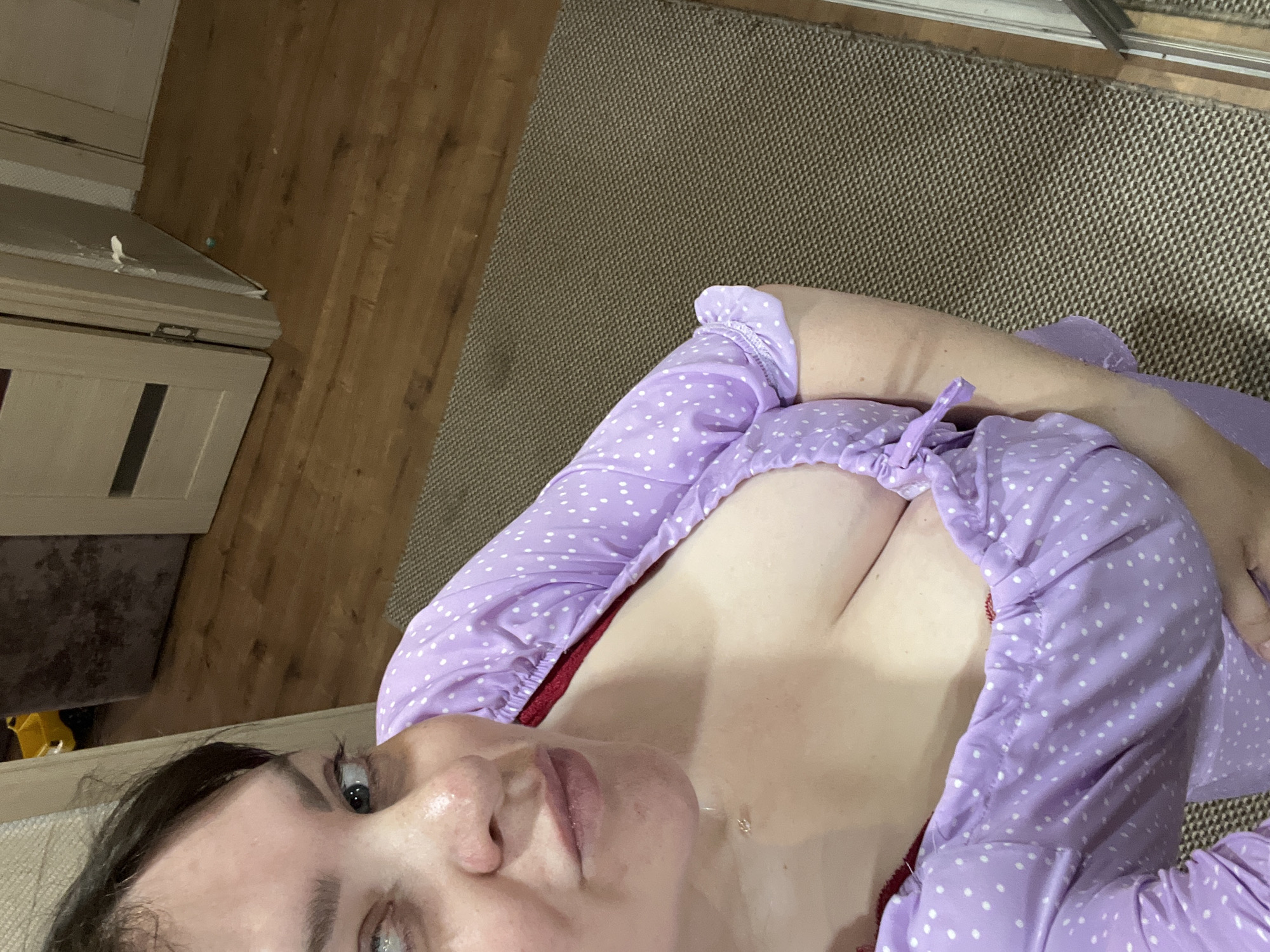 An adult woman is indoors, looking towards the camera with a slightly pursed expression. She is wearing a light purple, polka-dotted top with a low neckline, revealing part of her chest and a red bra. The background shows a wooden floor, a brown textured rug, and a partially open door with some white material protruding from its edge. A glimpse of a yellow toy is visible in the far background. The location is a residential interior in Noviy, Russia.