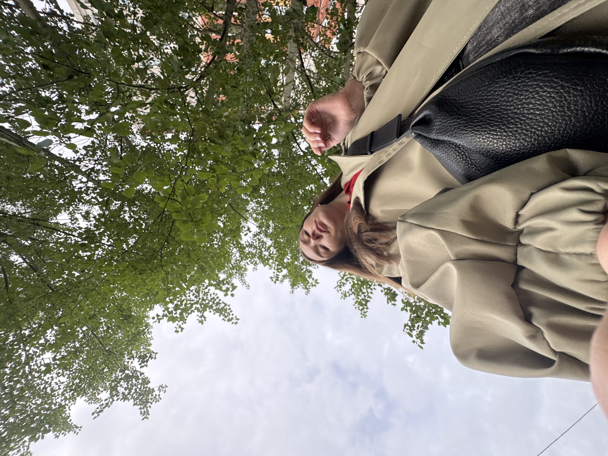 A low-angle shot looks up towards a woman standing underneath a tree, against a backdrop of a cloudy sky and tree foliage. The woman has brown hair and is wearing a beige trench coat with a black leather bag slung across her body. The location appears to be in a city with buildings partially visible behind the trees; however, the exact city of Izhevsk, Russia, is not verifiable from the image alone.