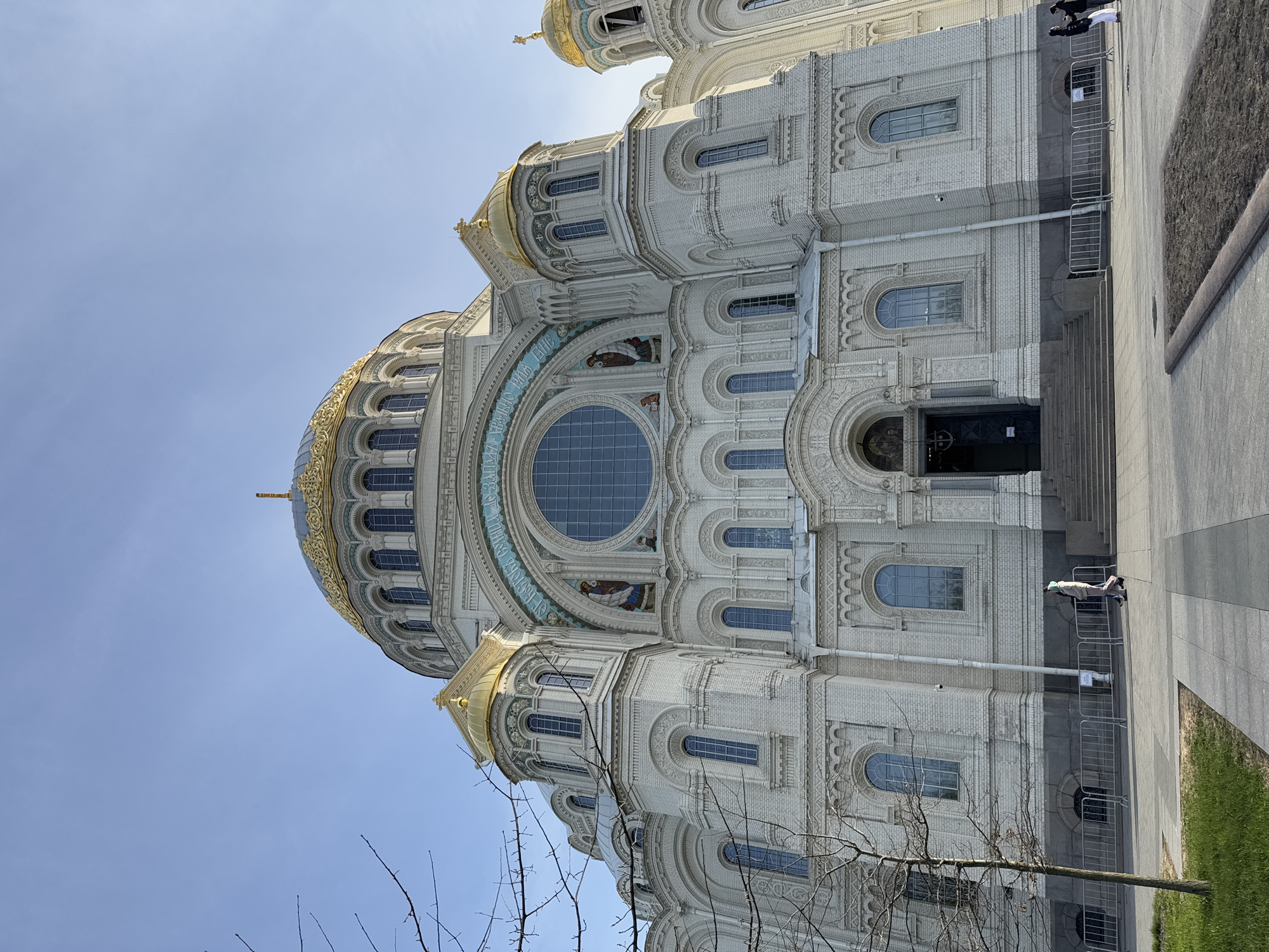 The image shows a partial view of the Naval Cathedral of Saint Nicholas in Kronstadt, Russia. The cathedral is predominantly white with gold domes. The architecture is ornate, featuring arched windows and detailed brickwork. A large, round window is a prominent feature of the facade. Two people are standing at the entrance, and some metal barriers are present. A bare tree with thin branches is visible in the foreground, and a patch of green grass is on the right side. The sky is light blue.