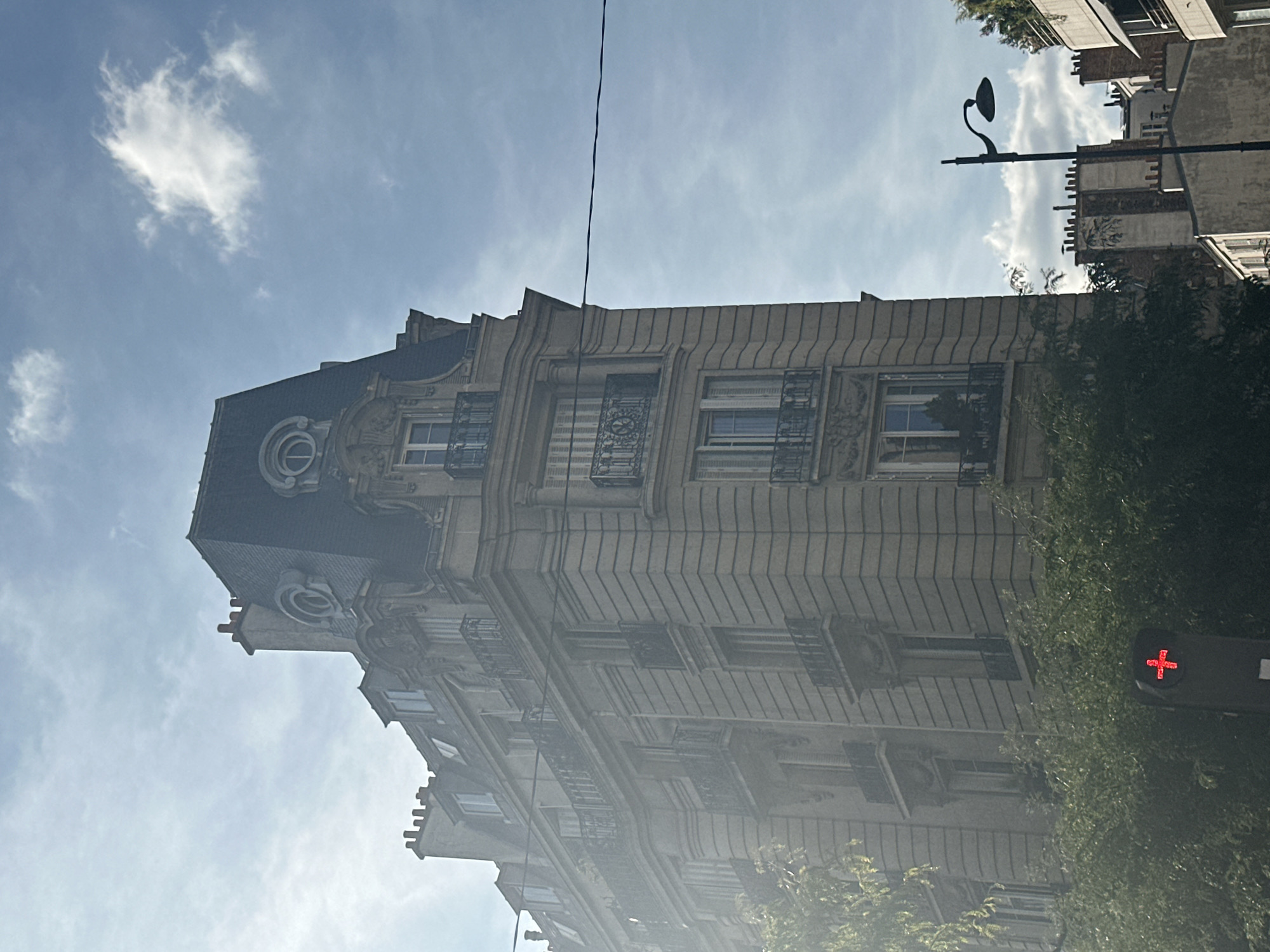 The image shows a Parisian apartment building, characteristic of Haussmannian architecture, on a sunny day with a partly cloudy sky. The building features ornate detailing, mansard roofs with dormer windows and chimneys, and wrought-iron balconies on several floors. A traffic light displaying a red cross is visible in the foreground, partially obscured by trees. A lamppost stands to the right of the building. The overall scene depicts a typical urban streetscape in Paris.