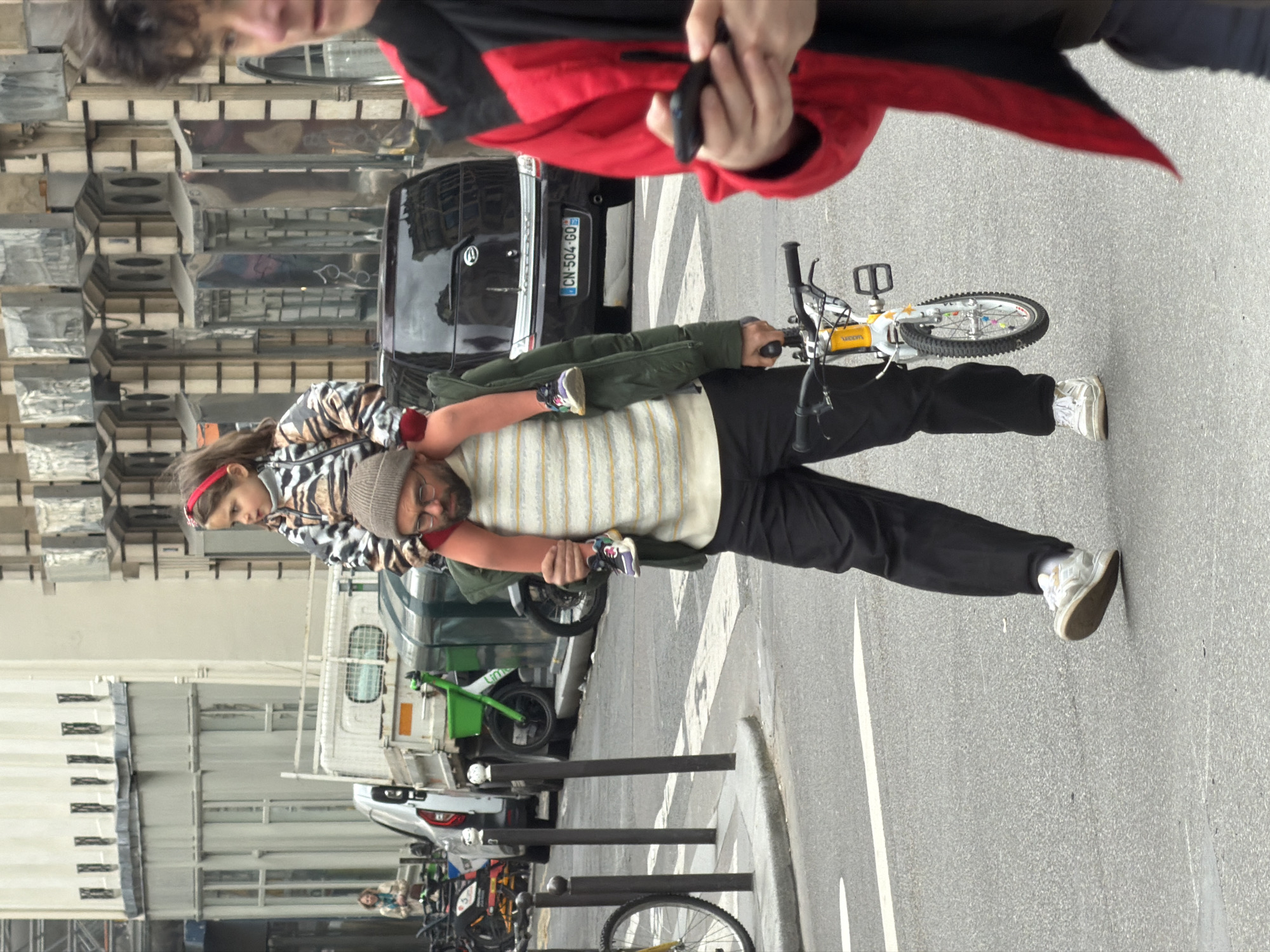 A medium shot shows a man with a beard and beanie walking on a Paris, France street with a small girl on his shoulders, carrying a bicycle. The man wears black pants, white sneakers, and a striped tan and white shirt. The girl is wearing a zebra-striped jacket and a red headband. The man is looking forward. The camera angle is slightly to the left. Behind the man is a parked black SUV with the license plate "CN-504-GO." Behind that, a person wearing a red jacket holds a phone. Street posts and parked cars and scooters are behind the man and to the left, with building facades in the background. A white stripe is on the ground, indicating a pedestrian crossing.