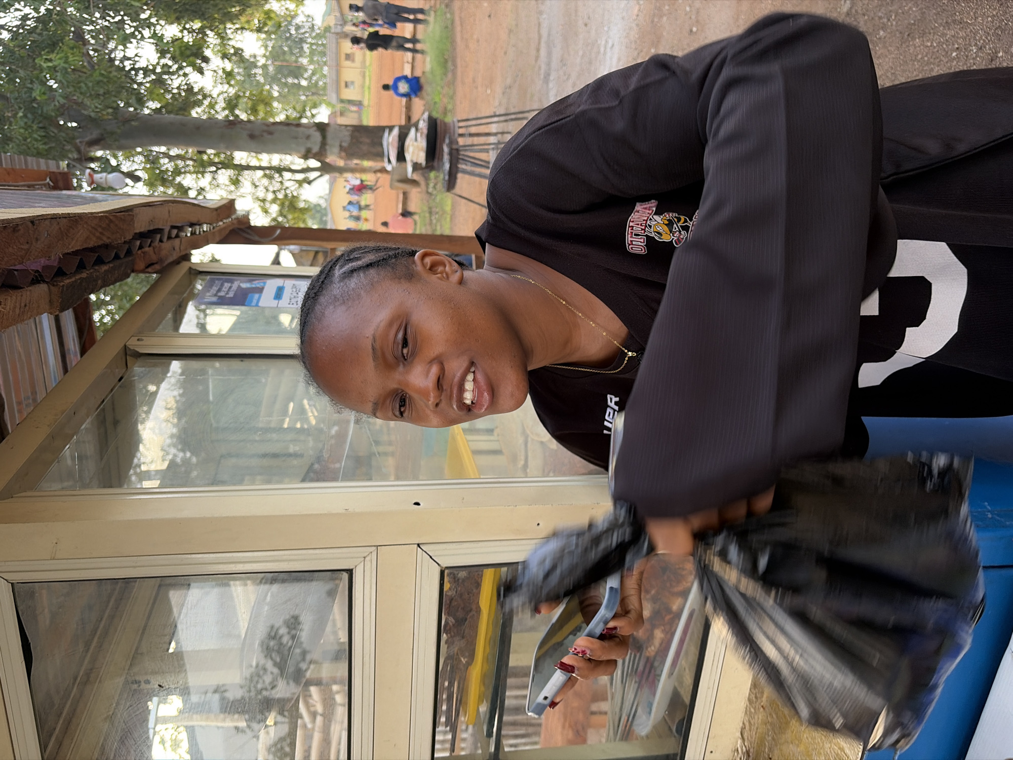 A young Black woman with braided hair stands in front of a glass-fronted food stall and smiles. She is wearing a black long-sleeved shirt with a "OTTAWA" logo and a cartoon bee on the left chest. Her right hand holds a smartphone, and her left hand clutches a black plastic bag. The stall has a corrugated metal roof and its glass panes reflect the surroundings. In the background, several people are visible, including a group of individuals standing near some buildings. The ground is reddish-brown dirt. The scene appears to be outdoors, possibly in a market or roadside setting in Rafin Par, Nigeria.
