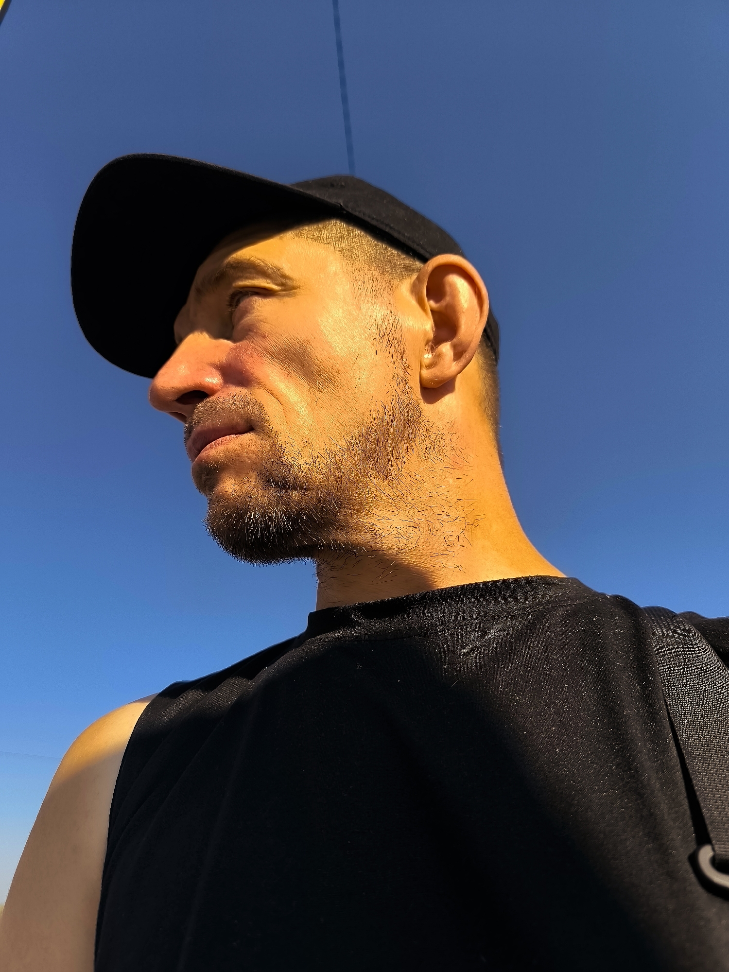 A low-angle, close-up, outdoor, profile shot shows a man wearing a black baseball cap and a black t-shirt against a bright blue sky. The man's face is illuminated by sunlight on the right side, casting a shadow on the left. He has stubble and is looking to the left. The background is clear blue sky with no visible elements of the city of Qarağandı.