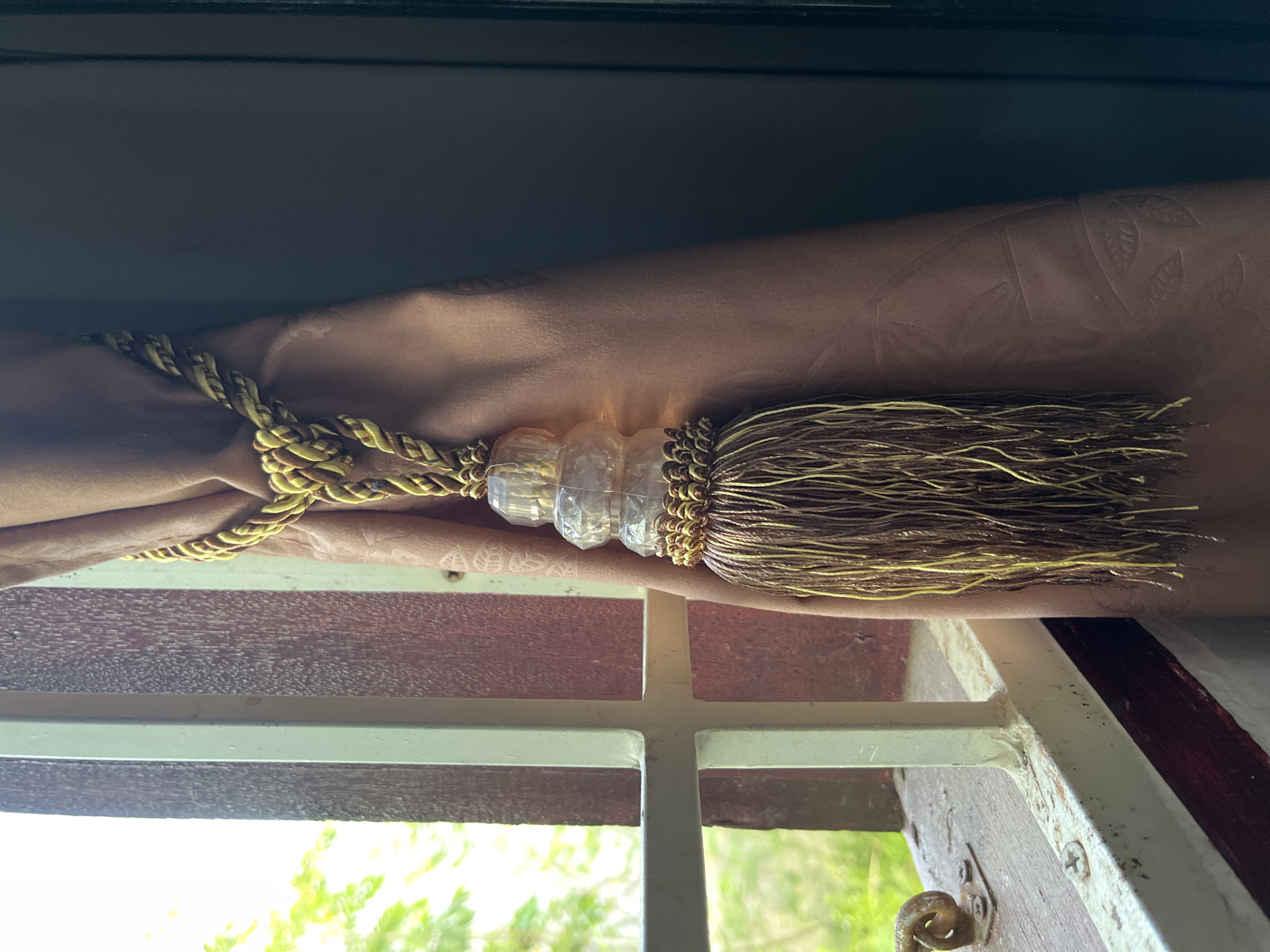 A tan curtain is held to the side of a window with a gold tassel tieback. The tassel has a decorative crystal-like band. The window is multi-paned with a white frame and a red-brown interior frame. Green foliage is visible outside the window. The scene appears to be indoors. No specific information indicates this is in Lhokseumawe, Indonesia.