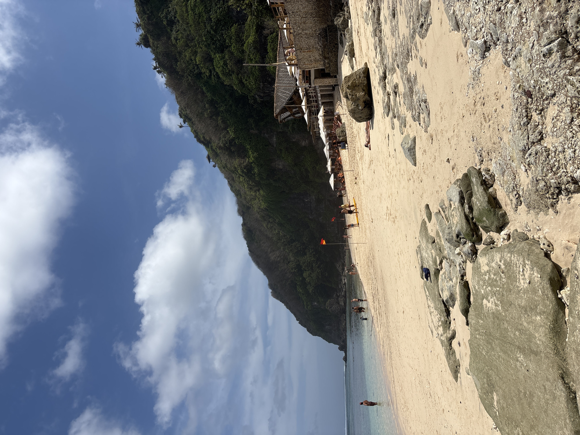 A wide, sandy beach in Kuta Selatan, Indonesia, stretches towards a lush, green hillside that dominates the background. The water is clear and shallow near the shore, with gentle waves lapping the sand. Several people are visible on the beach and in the water. Some are wading, while others are relaxing on the sand, some under umbrellas. A small beachside establishment with thatched roofs and white umbrellas is situated partway up the slope. Large rocks are scattered across the foreground of the beach. The sky is a vibrant blue with scattered white clouds. Two red flags are planted in the sand, possibly indicating swimming conditions.