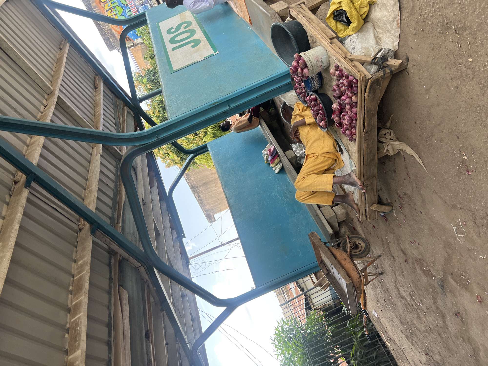 The scene shows an outdoor market stall in Jos, Nigeria, marked by a blue sign bearing the city's name. The stall is situated on a dirt ground surface with scattered debris. A person wearing yellow clothing is lying next to the stall. Various items are displayed for sale, including onions contained in buckets and bowls. A wooden cart stands beside the stall. A metal structure, potentially part of a canopy, is visible above the stall. The background features buildings, foliage, and power lines.