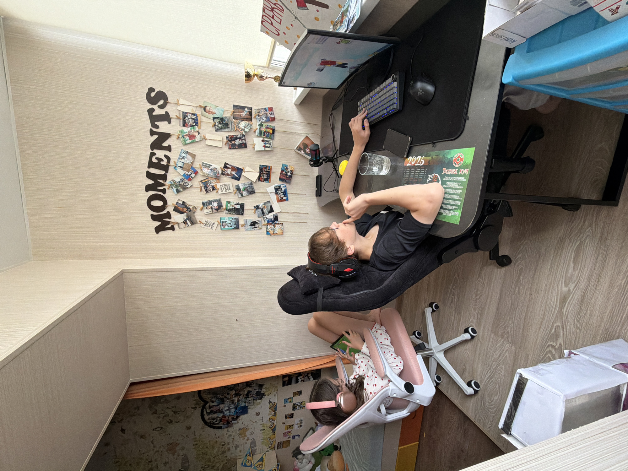 In a room with wood-patterned laminate flooring and cream-colored striped wallpaper, two individuals are engaged with electronic devices. On the left, a young girl with pink headphones sits in a pink and white gaming chair, looking at a tablet. She is wearing a white pajama top with red hearts. To the right, a person with glasses and headphones is seated in a black gaming chair, facing a computer monitor displaying a video game. Their hands are on the keyboard and mouse. A glass of water, a smartphone, and a mousepad are on the desk. Above the desk, the word "MOMENTS" is spelled out with wooden letters, and several small photographs are clipped to strings hanging on the wall. A trophy cup and a poster with red and white elements are also visible on the right side of the desk. The room appears to be a living space or bedroom, likely within a residential building in Birobidzhan, Russia.