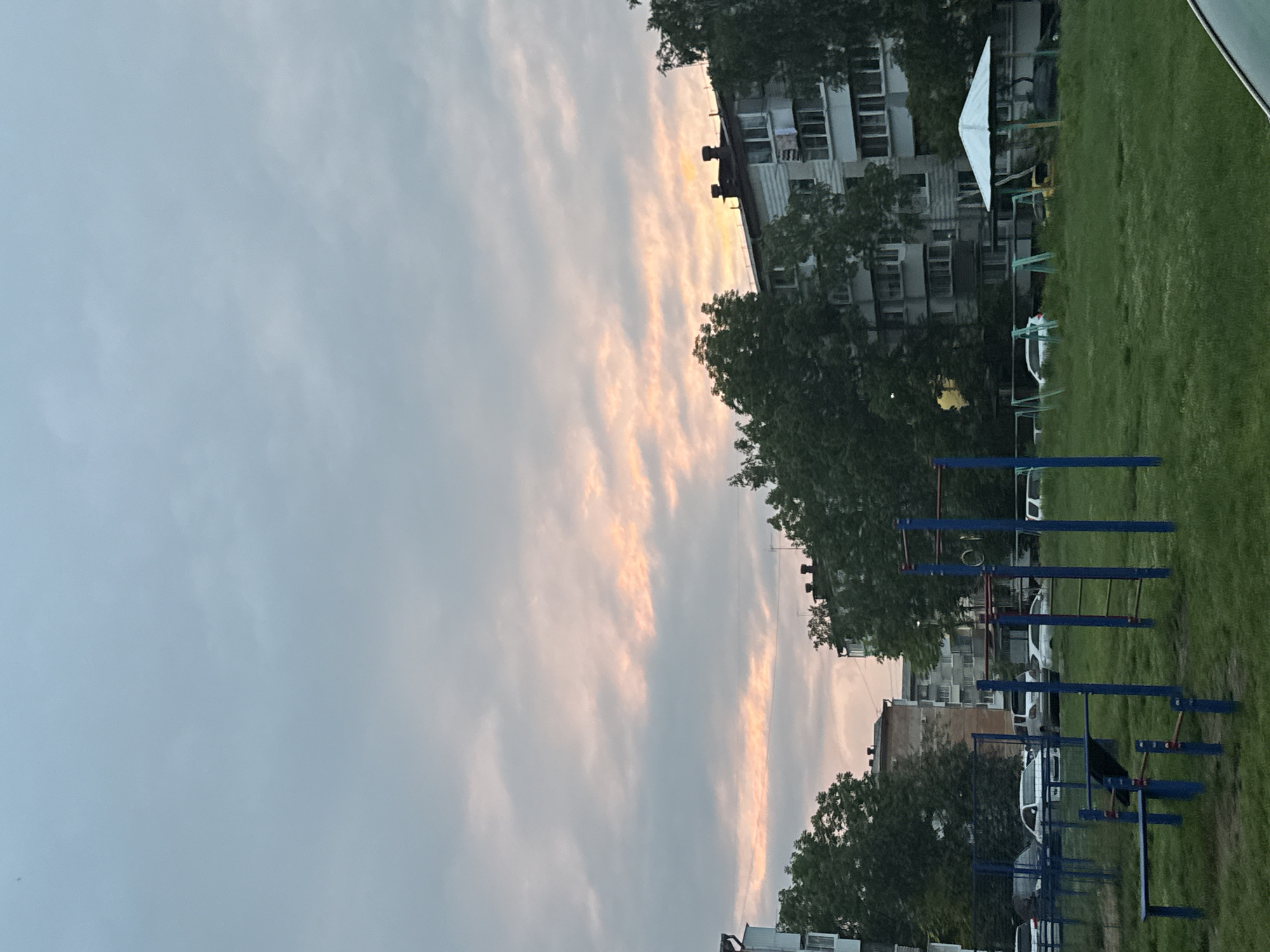 The image depicts an urban residential area in Birobidzhan, Russia, under a cloudy sky with patches of pinkish-orange light suggesting either dawn or dusk. In the foreground, a children's playground with blue metal bars and green turf is visible. Beyond the playground, there are parked cars and residential buildings, typical of a Soviet-era apartment complex. Trees line the area, partially obscuring the buildings and adding a touch of greenery to the urban landscape. The overall atmosphere is one of a quiet, everyday scene in a Russian city.