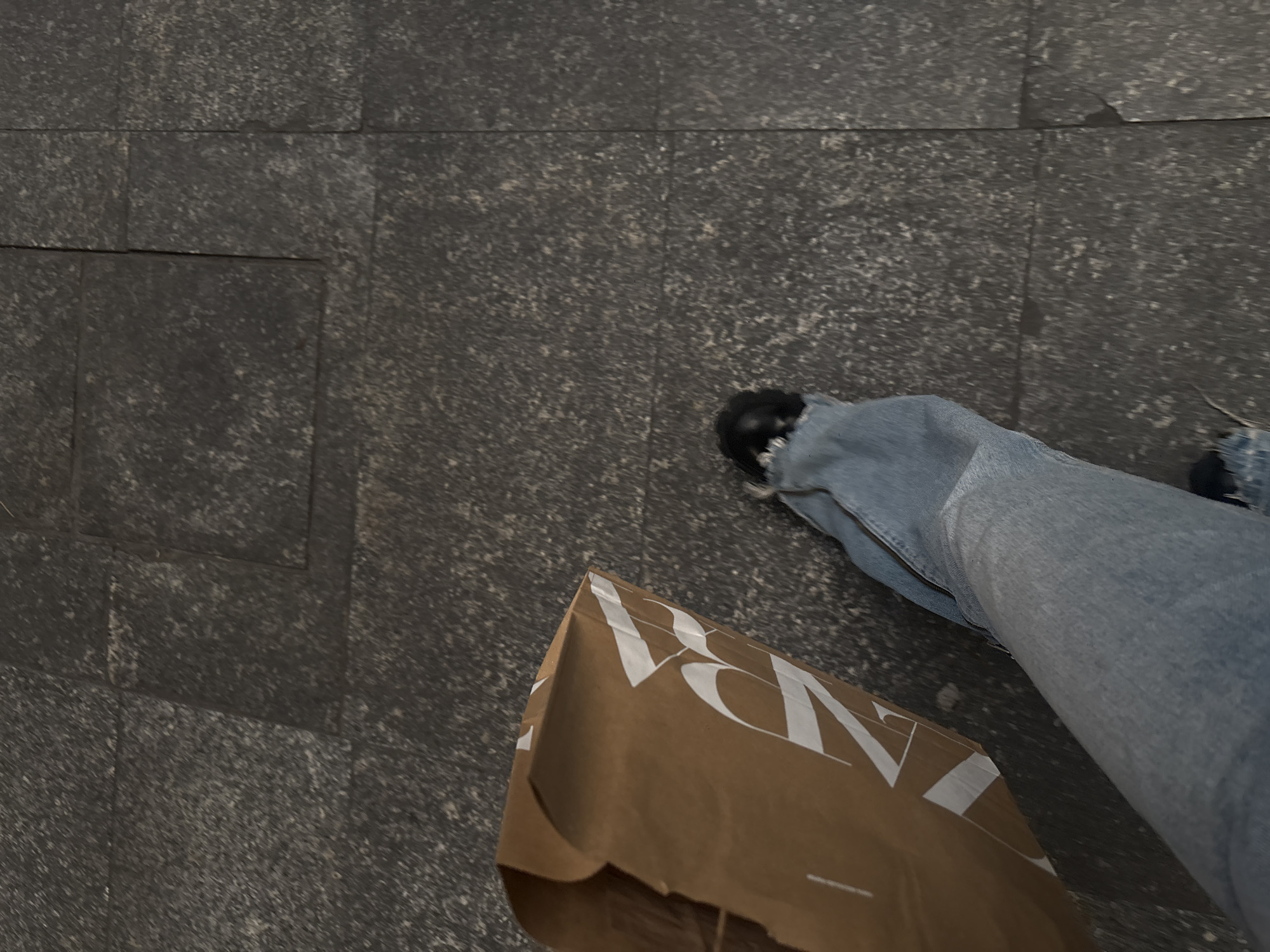 The image depicts a person walking on a gray-paved surface, likely a sidewalk. The lower right portion of the image shows a leg wearing blue jeans and black shoes. Next to the leg is a brown paper shopping bag with "ZARA" printed in white. The paved surface is composed of dark gray, textured tiles, some of which are square and others rectangular. The location is determined as Mexico City, Mexico, by the brand ZARA being a common shopping destination and the type of pavement being frequently used. The focus is on the person's progress with their recent shopping.