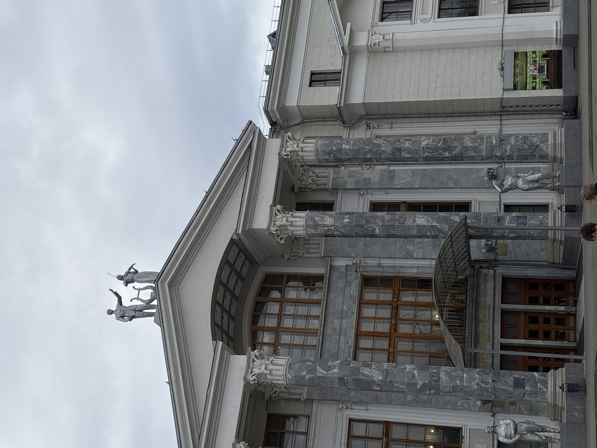 The image shows a low-angle view of the Perm State Art Gallery in Perm, Russia. The architectural style is classical, featuring a pediment topped with statues of a man and a woman, Corinthian columns, and large windows. The building's facade is made of white and gray stone. Two statues of figures appear to be suspended from the building's exterior. The sky is overcast. A sign is visible on the upper right corner of the building. A metal canopy is visible over the entrance. Part of a person's head is visible on the right side of the image.