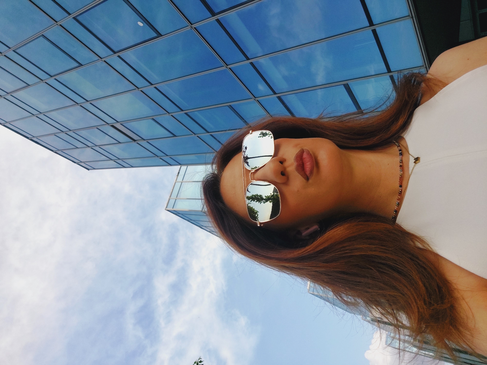 A woman with long, reddish-brown hair and sunglasses is standing in front of a modern glass building. The building appears to be made of glass panels and reflects the blue sky and some trees. The woman is wearing a white top and a necklace. The sunglasses have gold frames and reflect the surrounding environment. The image is taken from a low angle, looking up towards the woman and the building. The sky is partly cloudy. The location is identified as Sofia, Bulgaria, suggesting an urban setting.