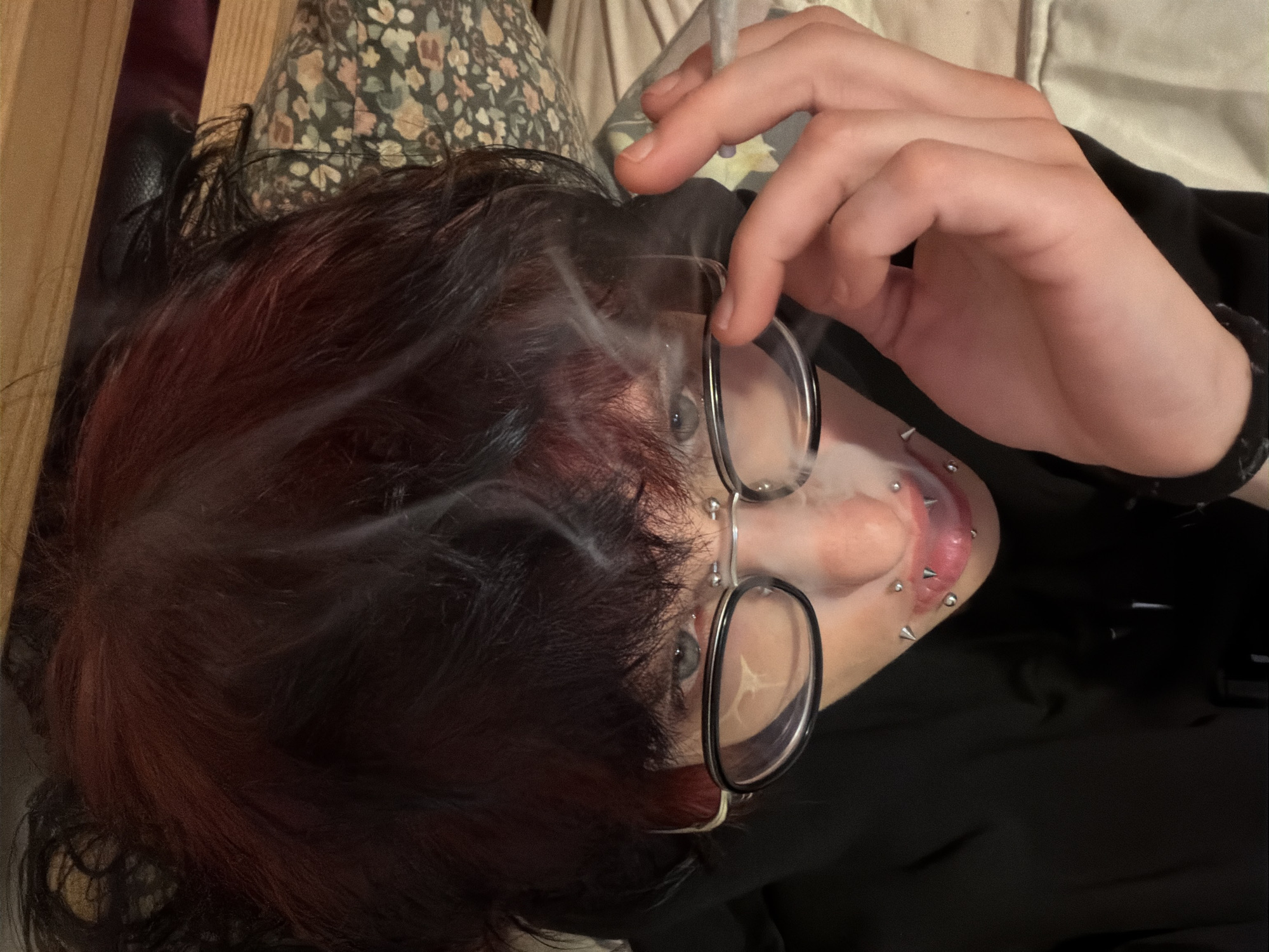 A person lies on a patterned pillow with black and dark red hair and glasses, holding a lit cigarette. The person's ear is visible and has several piercings. The person is wearing a black shirt. The location is not discernible from the image, and there is no indication it is in Sofia, Bulgaria.