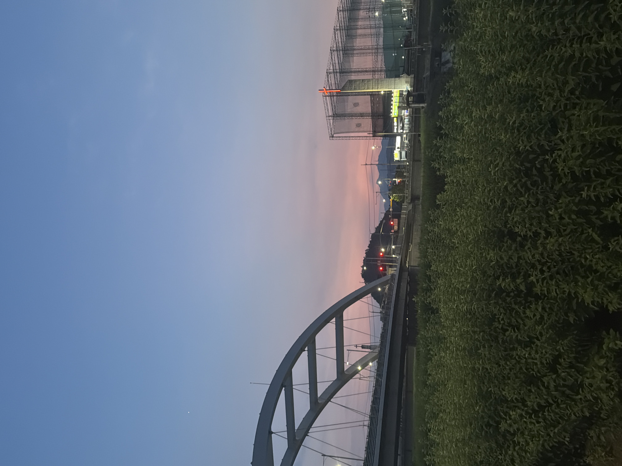 In Dongducheon-si, South Korea, at dusk, a picturesque scene unfolds. The sky transitions from a clear, pale blue at the top to a soft gradient of pink and orange near the horizon, with a faint star visible in the upper left. A prominent white arched bridge spans across a river or canal, its structural lines illuminated against the twilight. Below the bridge, a road carries a few vehicles, with traffic lights indicating a red signal. Across the river, a tall, light-colored building is topped with a bright red cross. This building is partially covered by scaffolding or netting, suggesting ongoing construction or maintenance. The surrounding area features lush green foliage in the foreground, obscuring the lower part of the scene, and distant hills or mountains form a silhouetted backdrop. Streetlights and building lights add to the ambiance of the setting.