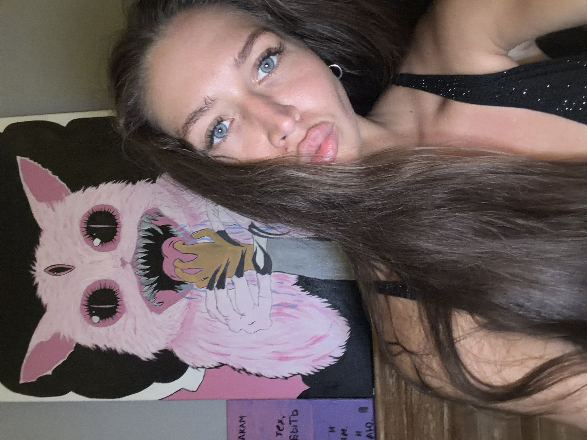 A woman with long brown hair and blue eyes is lying next to a painting of a three-eyed, pink, cartoon-like monster. The woman is wearing a dark, sparkly top and a small hoop earring. Her lips are slightly pursed. The painting has black background and some text in purple at the bottom. The scene is indoors. There is no indication in the image that this is in Canggu, Indonesia.