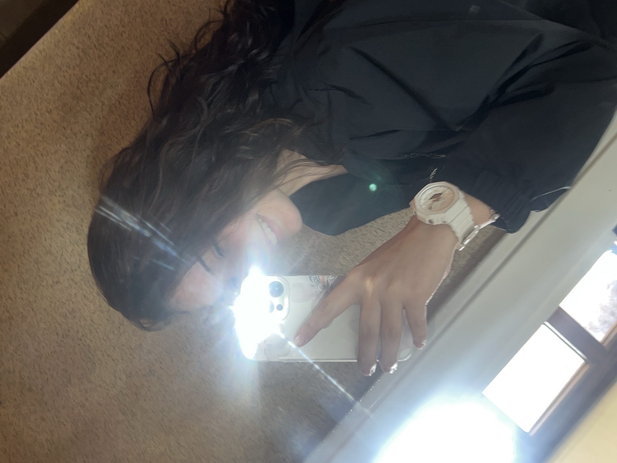 The image depicts an indoor mirror selfie of a young female, observed from a low angle. Her face is partially obscured by a bright lens flare originating from a light source in the lower left of the frame. She has dark, wavy hair and is smiling, with a portion of her teeth visible. She wears a black long-sleeved top or jacket. Her right hand holds a light-colored smartphone, possibly an iPhone, encased in a clear or light-colored case featuring a faint floral or butterfly pattern. The phone's camera module and a bright flash or strong reflection are prominent. On her left wrist, she wears a white or very light-colored wristwatch with an orange/pinkish analog-digital display. The background appears to be a light brown, textured wall or ceiling. A window frame, reflecting bright sunlight, is visible in the lower left. The specific location within the city of Elena, Bulgaria, cannot be determined from the visual information provided.