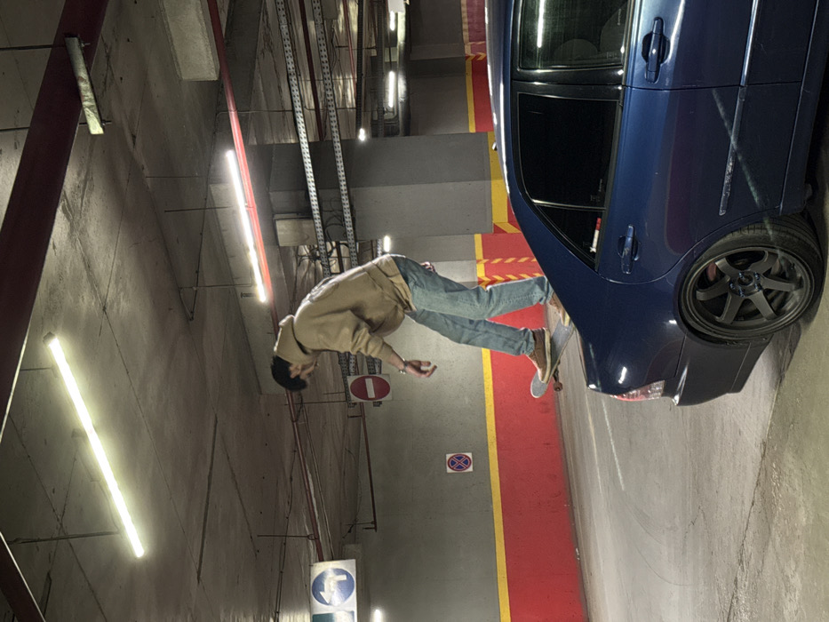 The scene depicts an underground parking garage in Sofia, Bulgaria, illuminated by fluorescent lights. A male individual, wearing a beige hooded sweatshirt, blue jeans, and brown shoes, is standing on a skateboard positioned on the trunk of a dark blue sedan. The vehicle features dark aftermarket multi-spoke wheels and tinted rear windows. The environment includes a concrete ceiling with exposed pipes and conduits, alongside concrete walls painted with a red upper section, a yellow horizontal stripe, and a grey lower section. Visible traffic signage includes a blue circular sign indicating a left turn, a red circular 'do not enter' sign, and a red circular 'no stopping/parking' sign. The individual's posture suggests an action of balancing or preparing for a skateboarding maneuver.