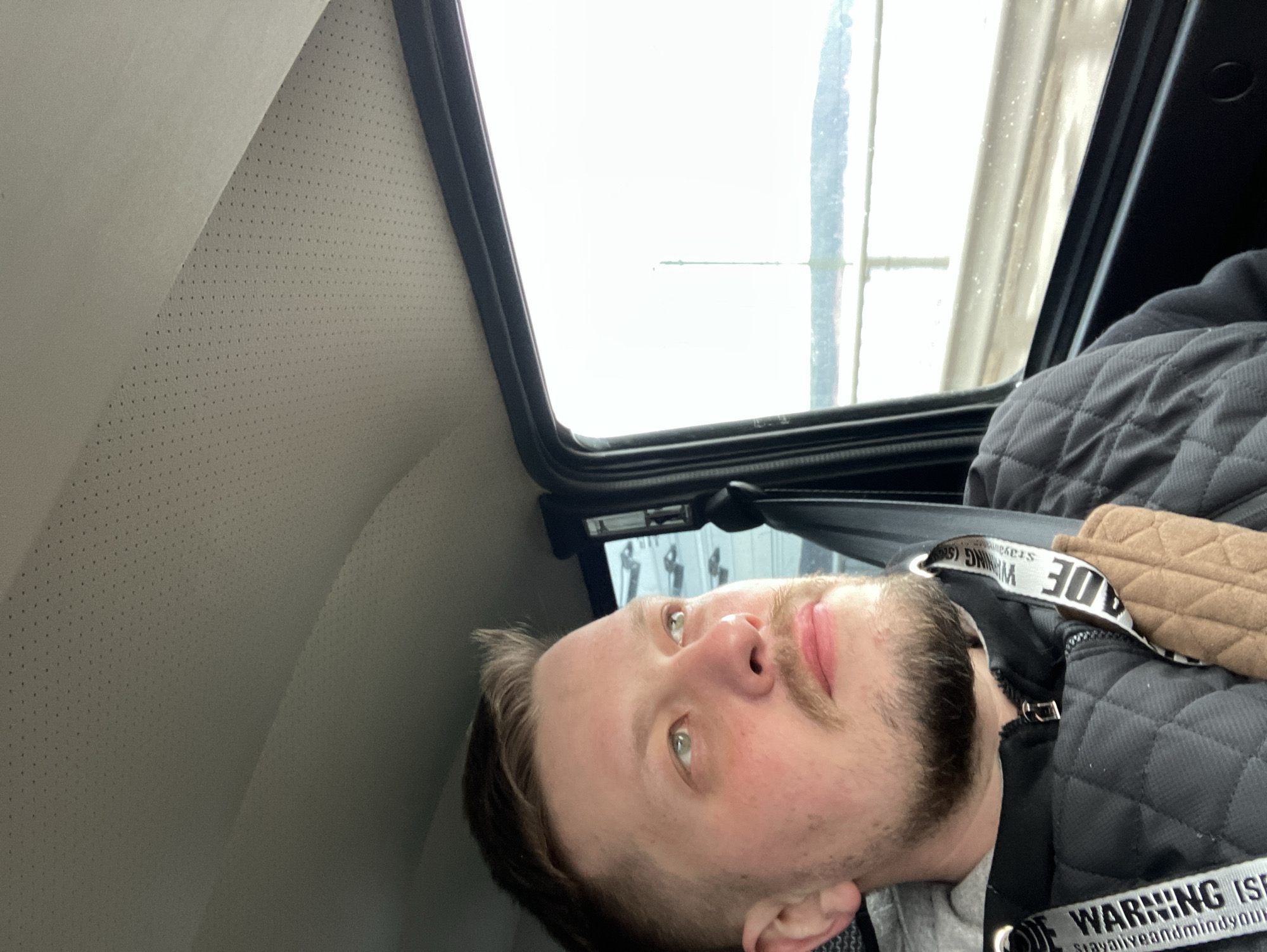 A man is lying down inside a vehicle, possibly a car or van. The interior features a light-colored, perforated headliner and a dark window frame. The man has fair skin, light eyes, and a beard. He is wearing a black quilted jacket with a tan shoulder strap. The strap has white text on it that reads "WARNING." Through the vehicle window, a blurry background is visible, possibly a building or structure. Based on the mention of the city of Chaikovsky, Russia, this image was likely taken there. However, without more visual context, it's impossible to pinpoint the exact location within the city.