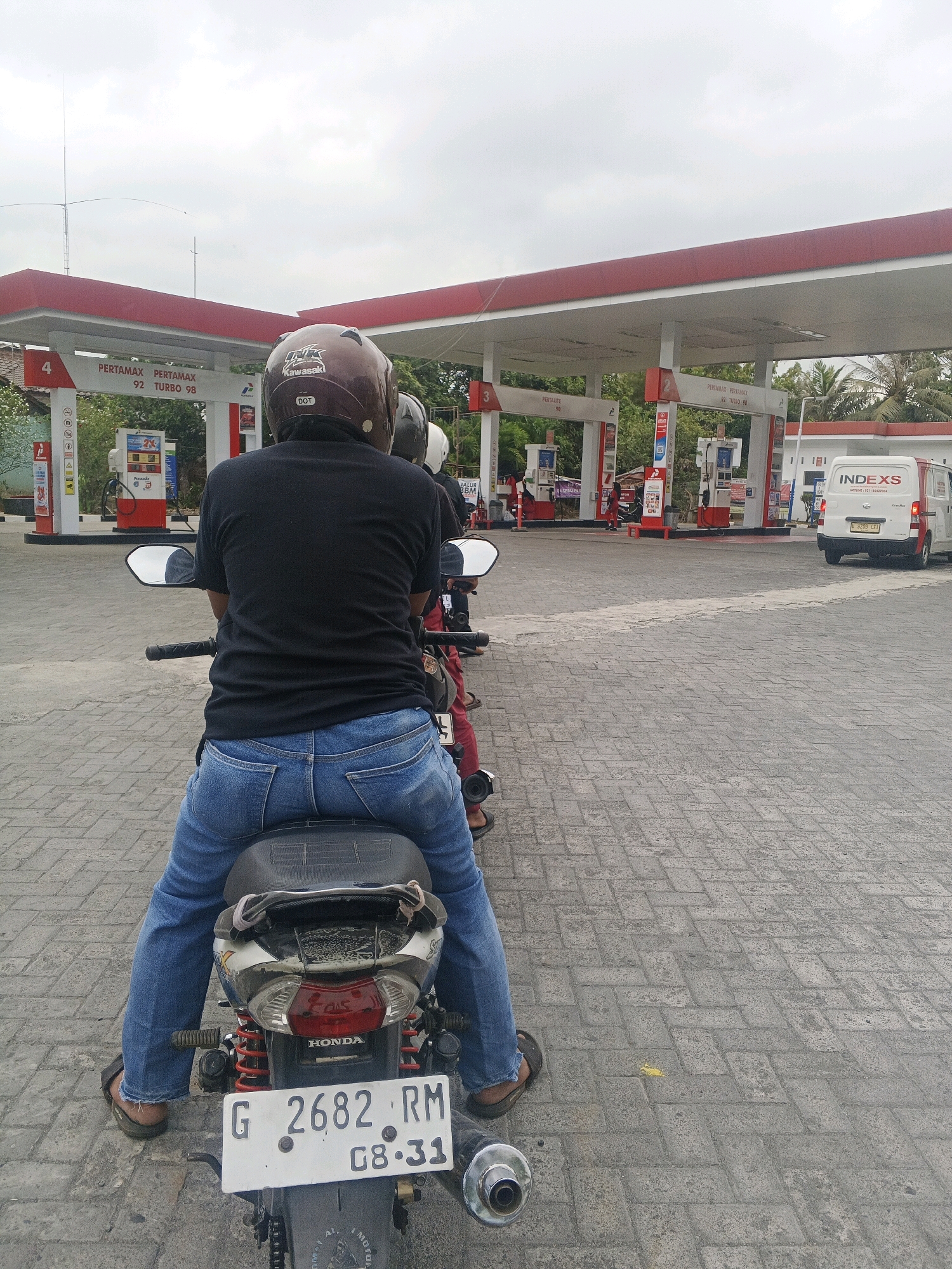 The image captures a scene at a gas station in Kliwonan, Indonesia, under an overcast sky. In the foreground, a person wearing a black t-shirt and blue jeans is seated on a motorcycle, facing away from the camera. The motorcycle has a license plate that reads "G 2682 RM 08-31". The person is wearing a brown helmet with "Kawasaki" and "DOT" visible. To the left of the main subject, another person is partially visible on a motorcycle.
The gas station features red and white canopies over gas pumps. Signs indicate "Pertamax 92 Turbo 98" and "Pertalite 90". Several gas pumps are visible, some with prices displayed. In the background, a white van with the "INDEXS" logo and a license plate "B 5209 CTT" is parked to the right. Trees and other structures are visible behind the gas station. The ground is paved with interlocking brick pavers.