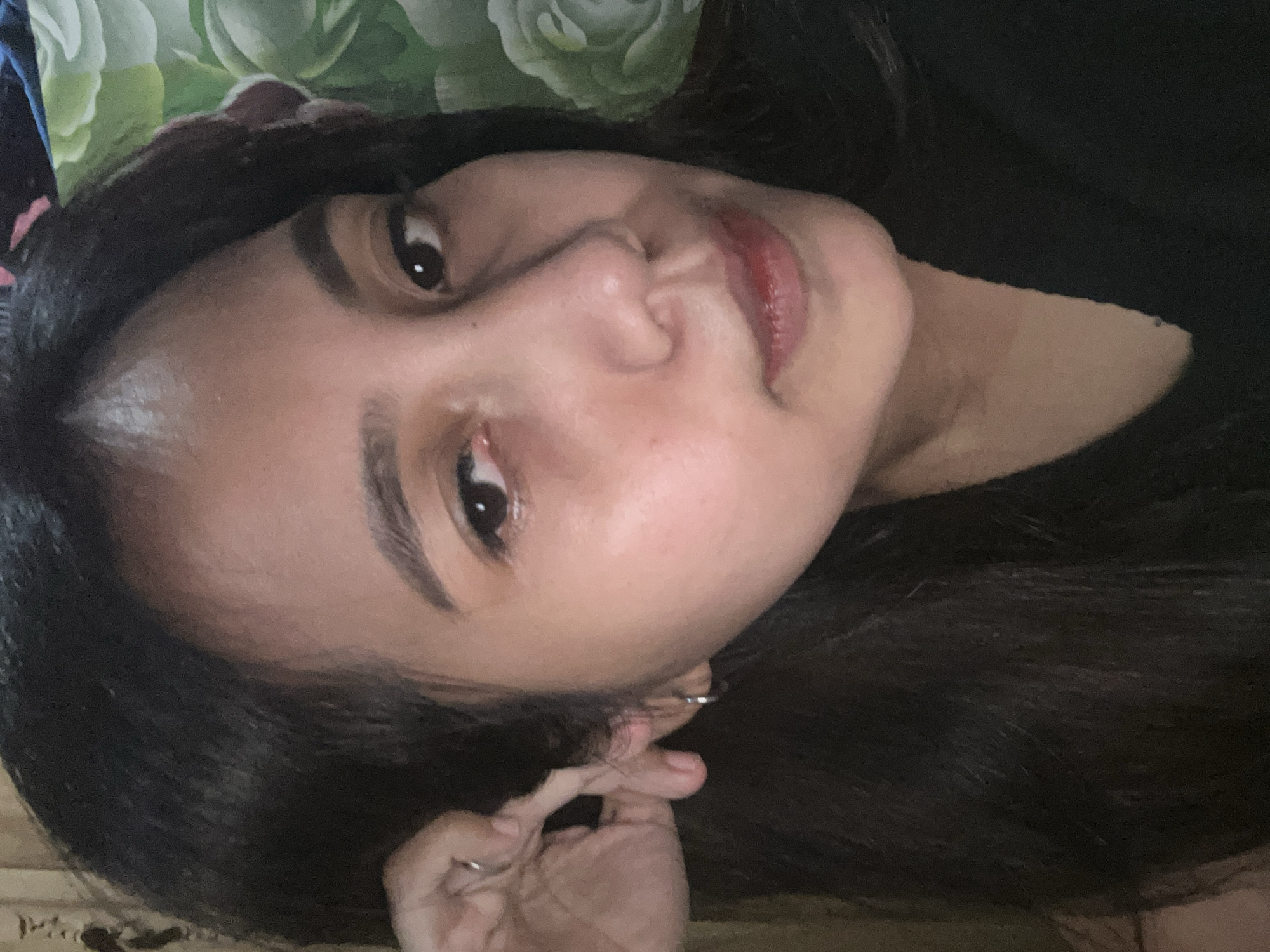 The image is a close-up, head-and-shoulders portrait of a young woman with dark hair. She is looking slightly to her left, with her right hand touching her ear. She is wearing a black top. The background appears to be a wall with a floral pattern. The lighting suggests an indoor setting, possibly a bedroom, in Depok, Indonesia.