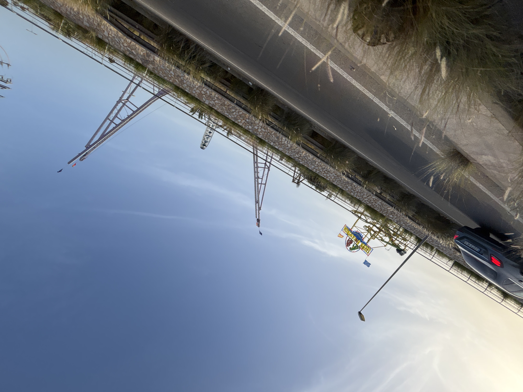 The image depicts a roadside view of what appears to be an amusement park or fairground located in the city of Orihuela, Spain, likely near the coast given the presence of a Ferris wheel and roller coaster-like structures against a clear blue sky. A dark-colored sedan is parked on the left side of the asphalt road, its brake lights illuminated. The road is lined with a stone wall, topped with vegetation and a railing, separating it from an elevated area that houses amusement park attractions. The Ferris wheel is visible in the background, and a roller coaster structure with tall, angled supports is prominent in the mid-ground. A sign with colorful lettering, possibly indicating the name of the amusement park, is also visible. The sun is low in the sky, casting a warm light and long shadows, suggesting it is either early morning or late afternoon. The overall scene is one of an urban entertainment district on a bright, clear day.