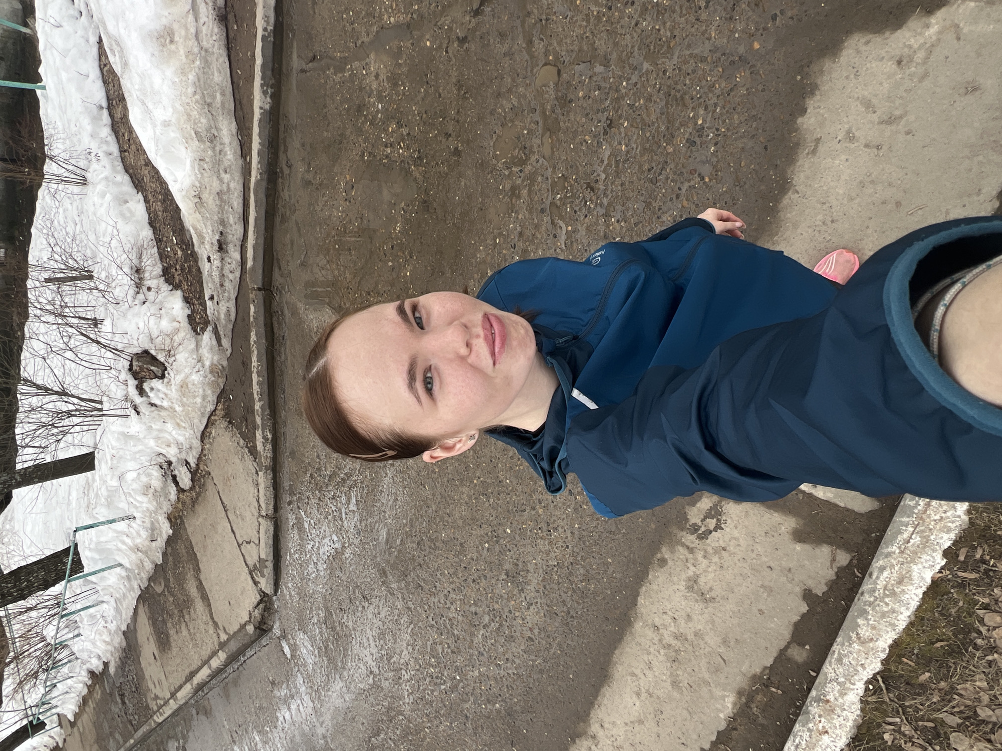 A young woman is taking a selfie outdoors in what appears to be a residential area in Chaikovsky, Russia. She is wearing a blue jacket. A sidewalk and an area with melting snow and bare trees are visible on the left. The ground is a mix of concrete and wet asphalt. A concrete border can be seen on the lower right side of the image with grass/bare foliage near it.