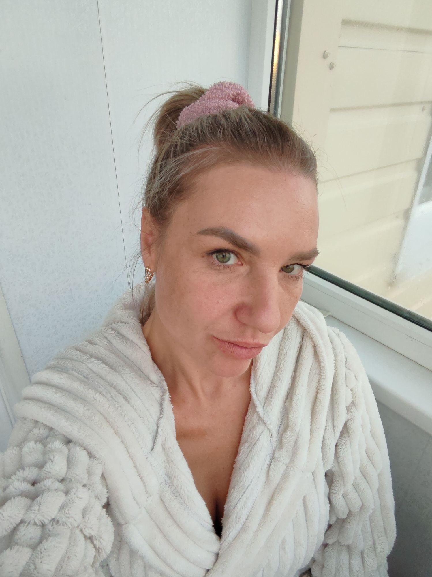 A woman is taking a selfie indoors, wearing a plush white bathrobe and a pink scrunchie in her hair. She has light blonde hair pulled back in a bun and green eyes. She is wearing gold earrings. The background appears to be a window with white blinds or paneling. The image is taken from a slightly high angle, looking down at the woman. The lighting suggests it is daytime, possibly near a window with natural light. There are no other people or objects clearly visible in the frame besides the woman and her immediate surroundings. The overall impression is of a casual, personal moment captured in a private indoor space. The location is not specified in the image.