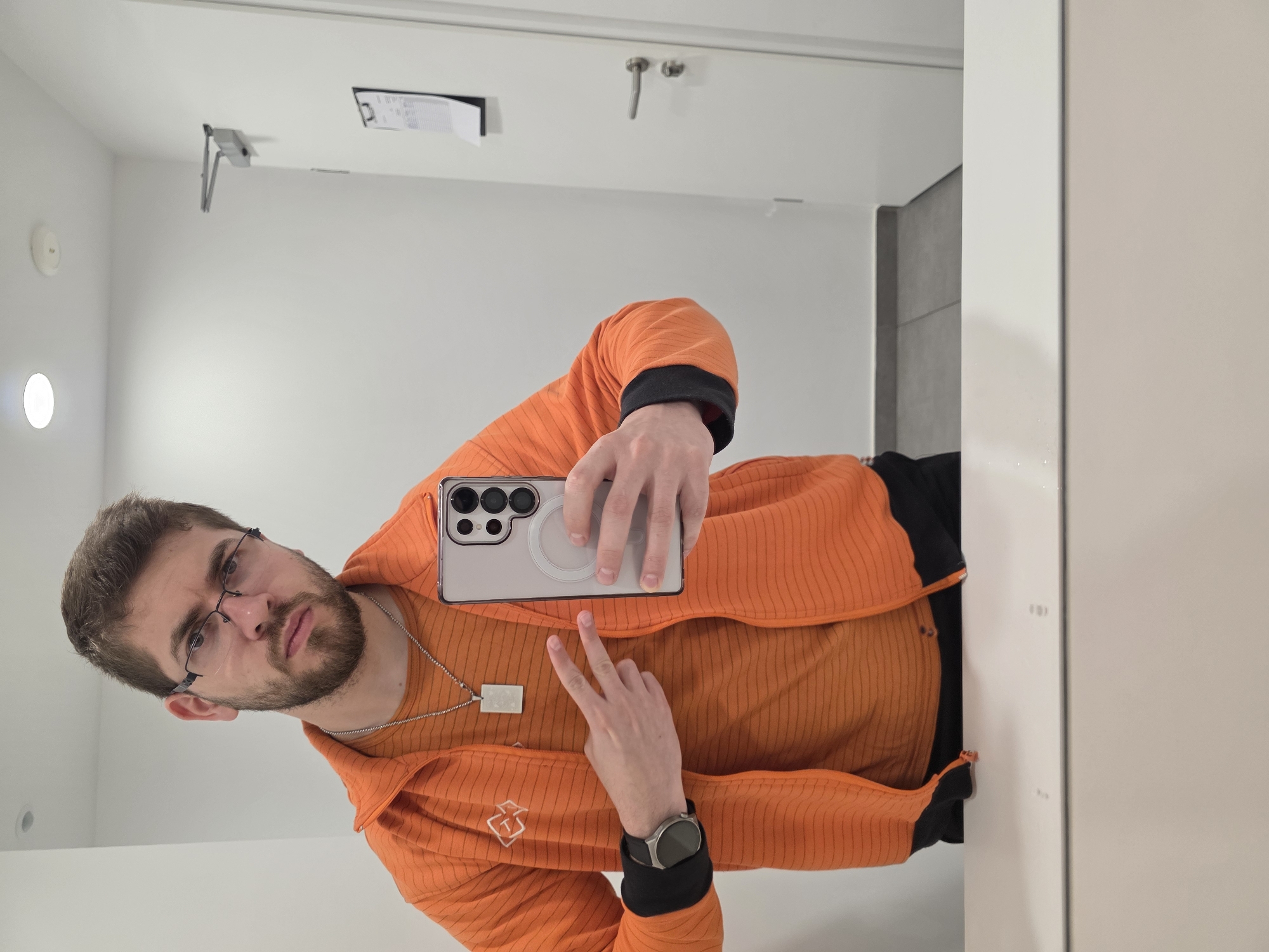 A man with brown hair, a beard, and glasses is taking a selfie in a bathroom or toilet. He is wearing an orange jacket, an orange shirt, a silver necklace, and a smartwatch. He is holding a cell phone with a clear case in his right hand, and he is making a peace sign with his left hand. The location appears to be in Plovdiv, Bulgaria, based on the presence of signage in Bulgarian Cyrillic in the background, although specific landmarks or characteristics that confirm the location cannot be identified in the immediate environment. The bathroom features white walls, a grey tiled area and standard fixtures.