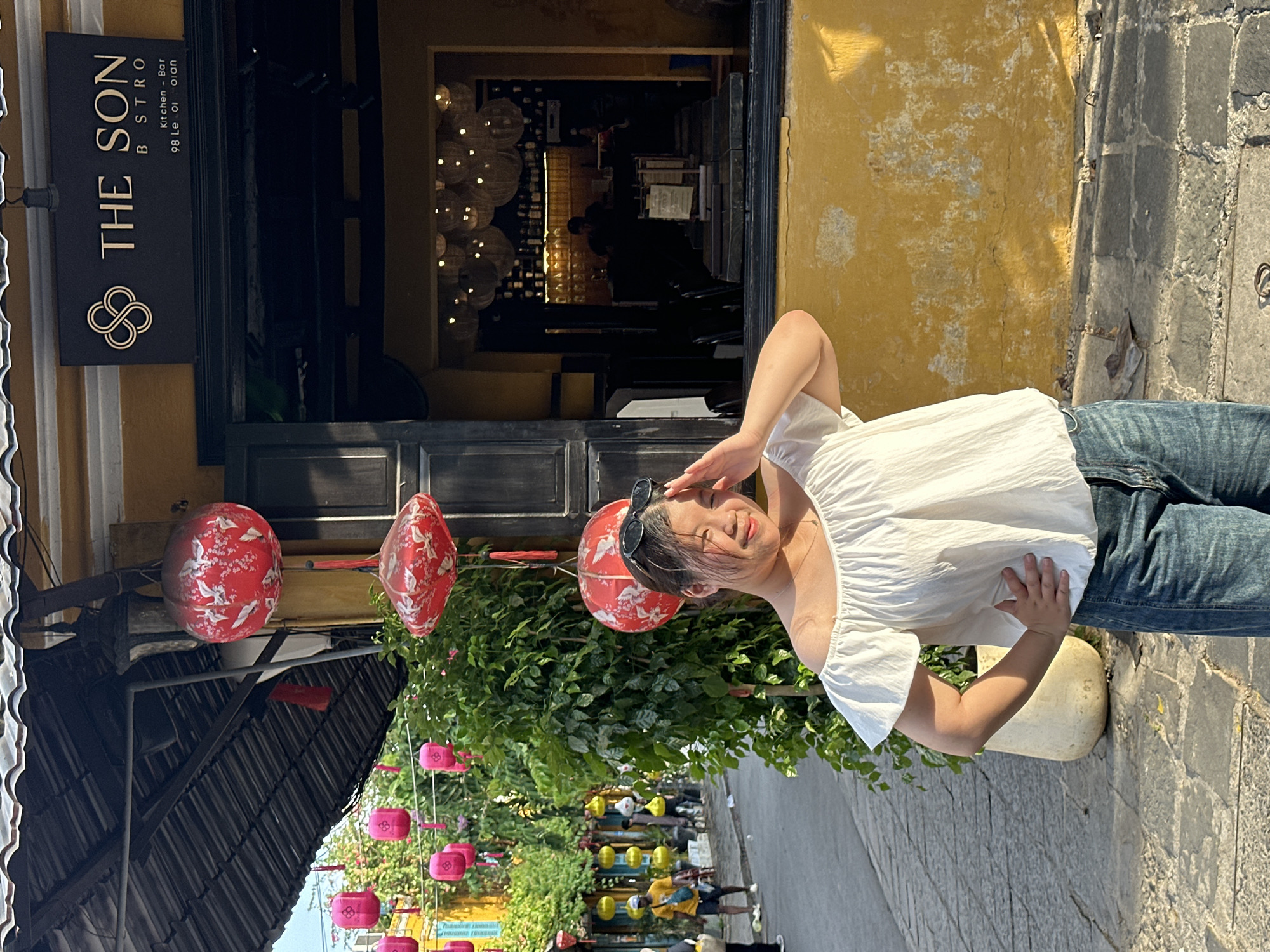 A young East Asian woman with dark hair pulled back, wearing sunglasses perched on her head, poses in front of a yellow building with a dark sign that reads "THE SON BISTRO Kitchen - Bar 98 Le Loi street". She is wearing an off-the-shoulder white top and blue jeans. Her right hand is raised to her forehead as if shielding her eyes from the sun, and her left hand rests on her hip. Behind her, the street is lined with green trees and decorated with red and pink lanterns. The street pavement is made of cobblestones. In the background, other people can be seen walking and sitting. The building facade is yellow and weathered.