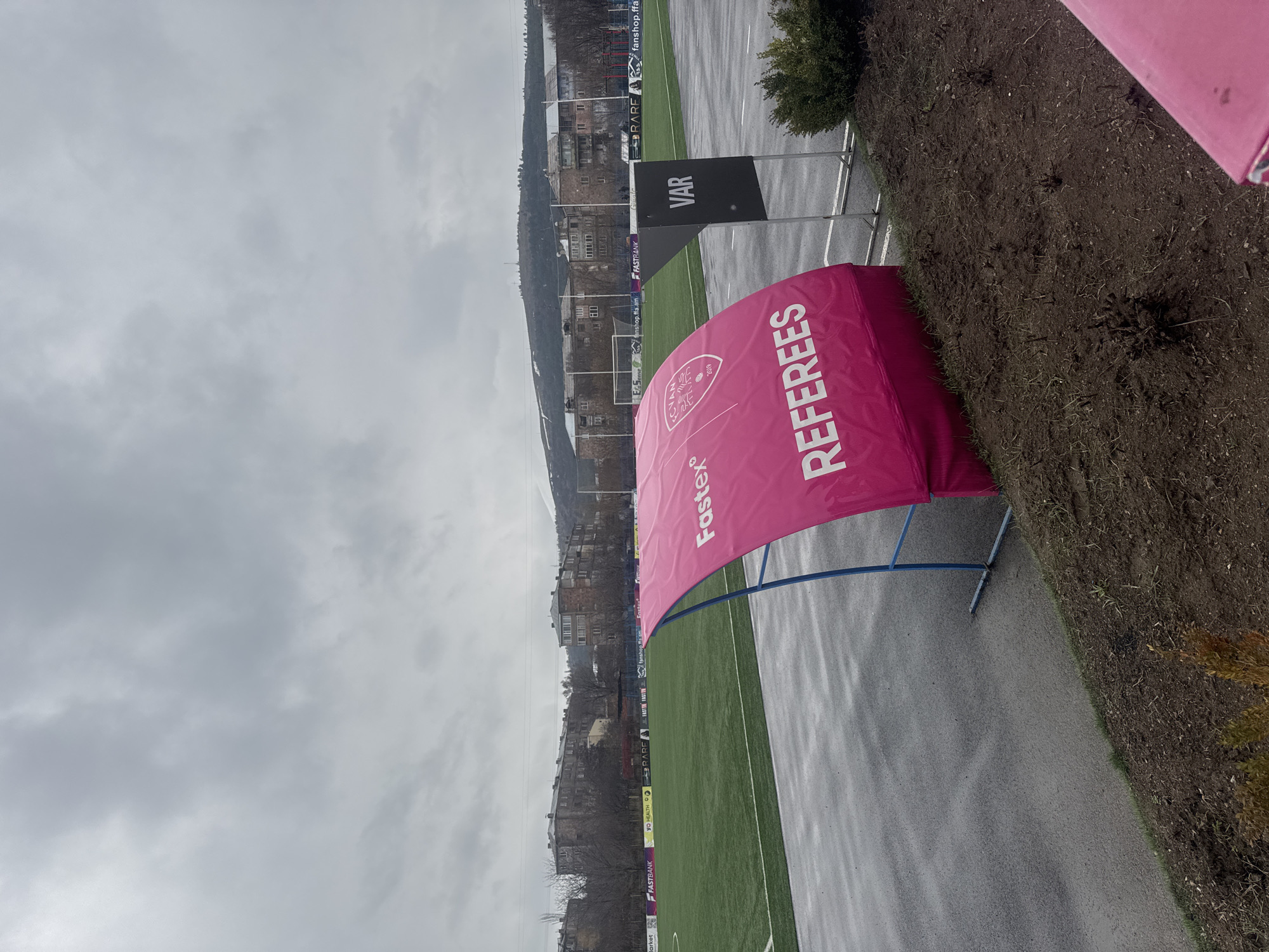 The image shows a view of a soccer field in Charentsavan, Armenia, on a cloudy day. A pink "REFEREES" tent is visible near the edge of the field, along with a sign that says "VAR." The background includes buildings and hills, typical of the area. The field appears to be artificial turf, and a wet pathway runs alongside it. Sponsorship banners line the field, with visible logos including "FASTBANK" and "YO HEALTH." The overall impression is of a match day setup, possibly during inclement weather.