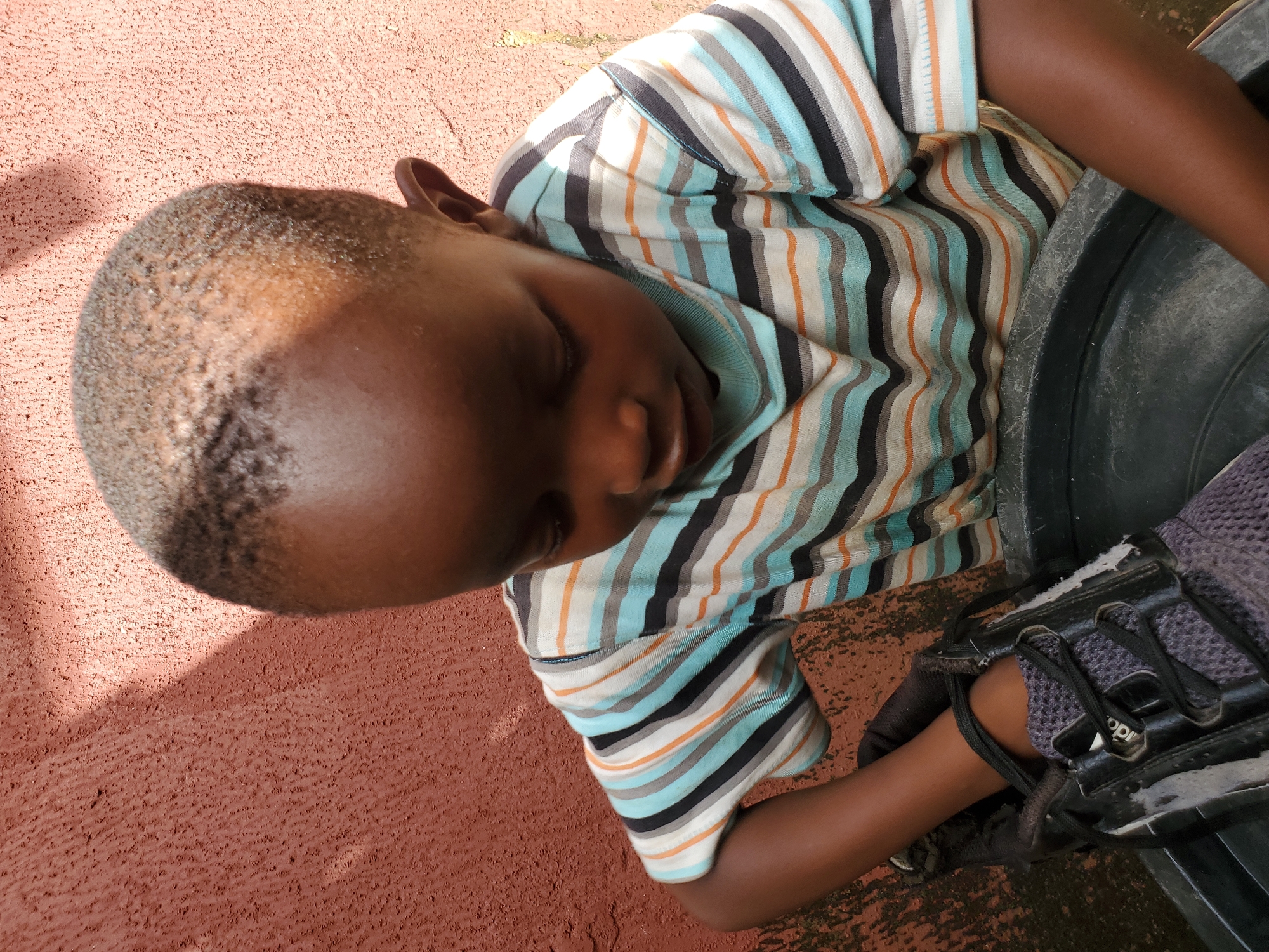 In an outdoor setting with a textured, reddish-brown wall to the left and right, a young Black child is captured in a close-up, overhead shot. The child is wearing a horizontally striped t-shirt with bands of white, light blue, orange, and black. The child's gaze is directed downwards, and their head is tilted to the right. A dark-colored shoe with grey fabric and black laces is positioned on the left side of the frame, with the child's right arm and hand partially inside it. The child is also leaning over a large, dark grey plastic basin. The scene appears to be in Jos, Nigeria, suggested by the context of the location.