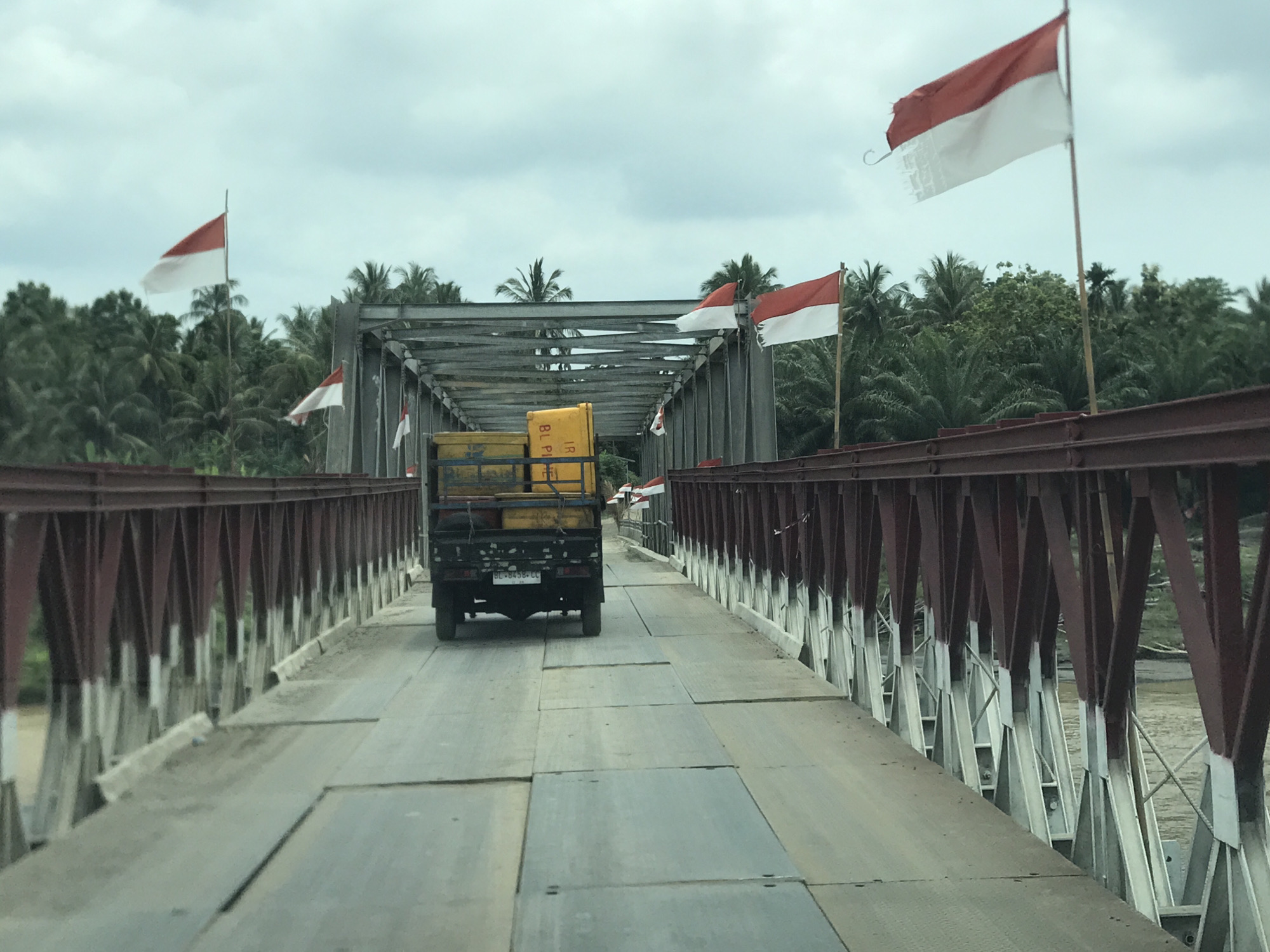 A black pickup truck, seen from behind, drives along a metal truss bridge in Teupinreudeup, Indonesia. The truck carries several bright yellow containers or crates, with the visible license plate reading "BL 8458 LL". The bridge features reddish-brown painted steel girders and a road surface constructed from large, somewhat uneven metal or wooden planks.
Numerous red and white Indonesian flags are displayed prominently along both sides of the bridge, attached to poles and the bridge structure itself. Some flags appear worn or tattered, with frayed edges, particularly a large one visible on the left side. The sky is overcast with light grey clouds, suggesting a cloudy daytime.
Beyond the bridge, dense tropical vegetation, primarily palm trees, covers the landscape, indicating a lush, possibly rural or coastal setting. A hint of brown water or a river is visible beneath the bridge on the left. The scene captures a moment of travel over an essential piece of local infrastructure, adorned with national symbols.