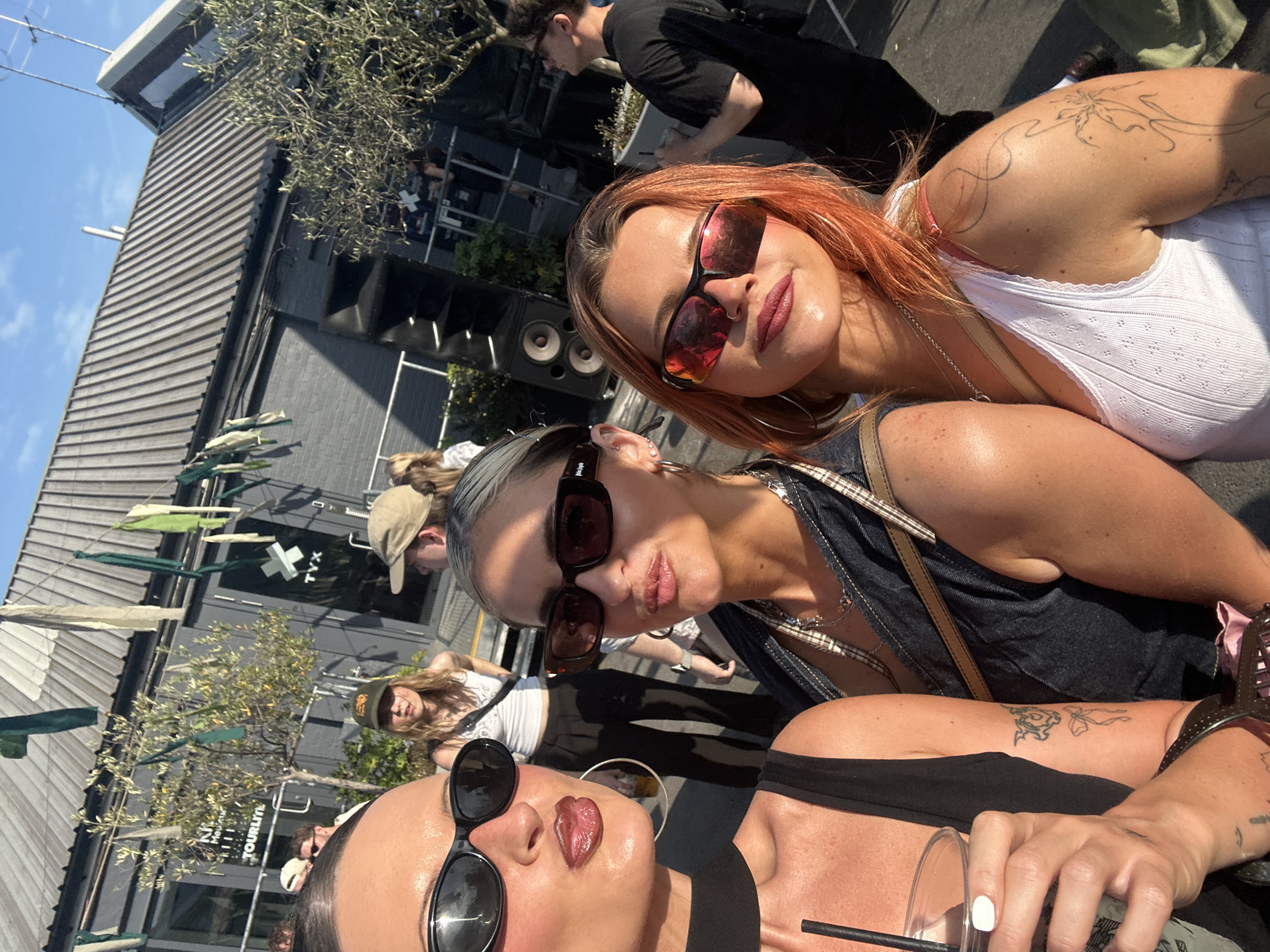The image is a selfie taken at an outdoor event in London, United Kingdom. Three women are the primary subjects, posed closely together. The woman on the top right has red hair and is wearing sunglasses and a white tank top, with a tattoo visible on her upper arm. The woman in the middle has light hair and is wearing sunglasses and a dark denim top with a cross body bag. The woman at the bottom is wearing sunglasses and a black top, holding a clear plastic cup with a straw and nail polish on her fingernails.
In the background, blurred figures suggest a crowd. A dark building with speakers and signage is visible, along with green and white hanging decorations. The sky is blue, indicating a sunny day. The sign on the building says TYX. The lower portion of the sign says "Kiki House Tourlife."