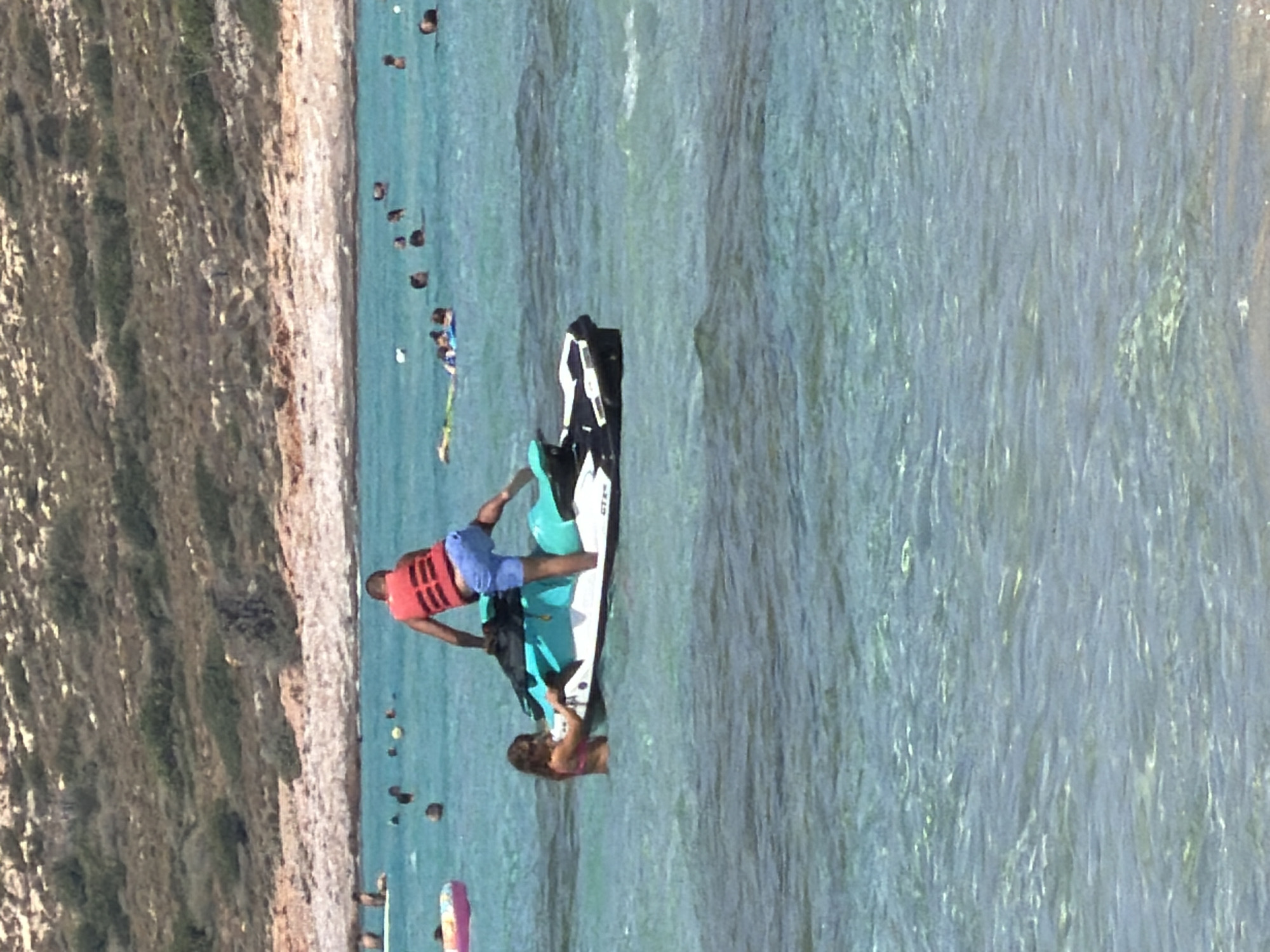 In the waters of an unknown location in Greece, a man wearing a red life vest and blue shorts is standing on a jet ski. Next to the jet ski, a woman in a pink bikini is partially submerged in the water, holding onto the jet ski. In the background, other people are swimming and floating in the ocean, with a sandy beach and a hilly, vegetated landscape visible on the shore. The water is clear and turquoise, with sunlight reflecting off its surface.