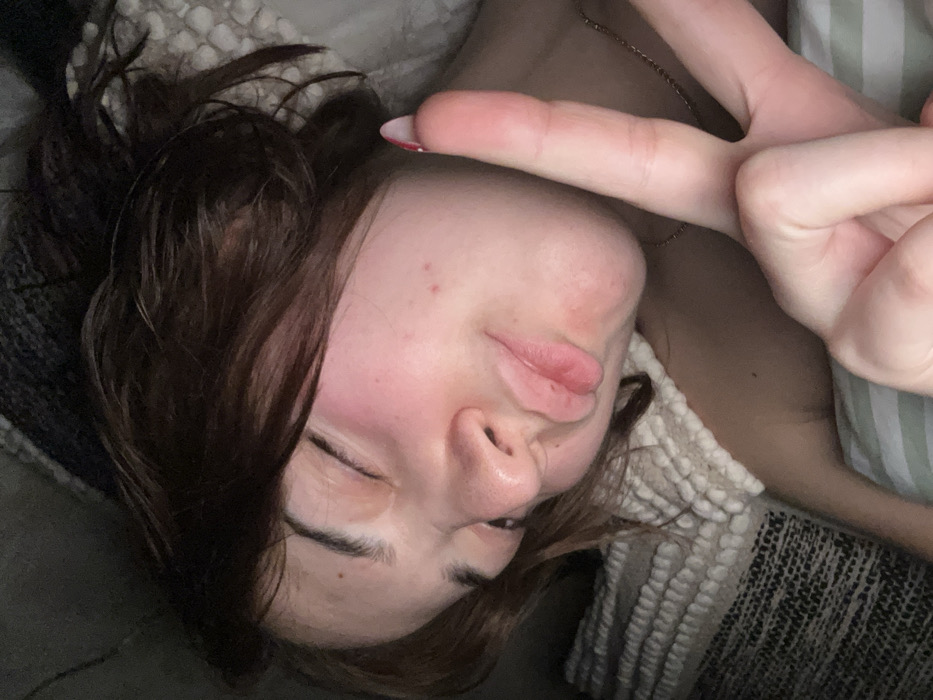 A woman lies sideways on soft bedding with dark brown hair framing her face. Her eyes are closed, and she makes a duck-like pouting expression with her lips. Her hand is raised above her face, forming a peace sign with her index and middle fingers. A delicate gold chain necklace is visible. The background consists of layered blankets with varying textures, including a thick knitted one.