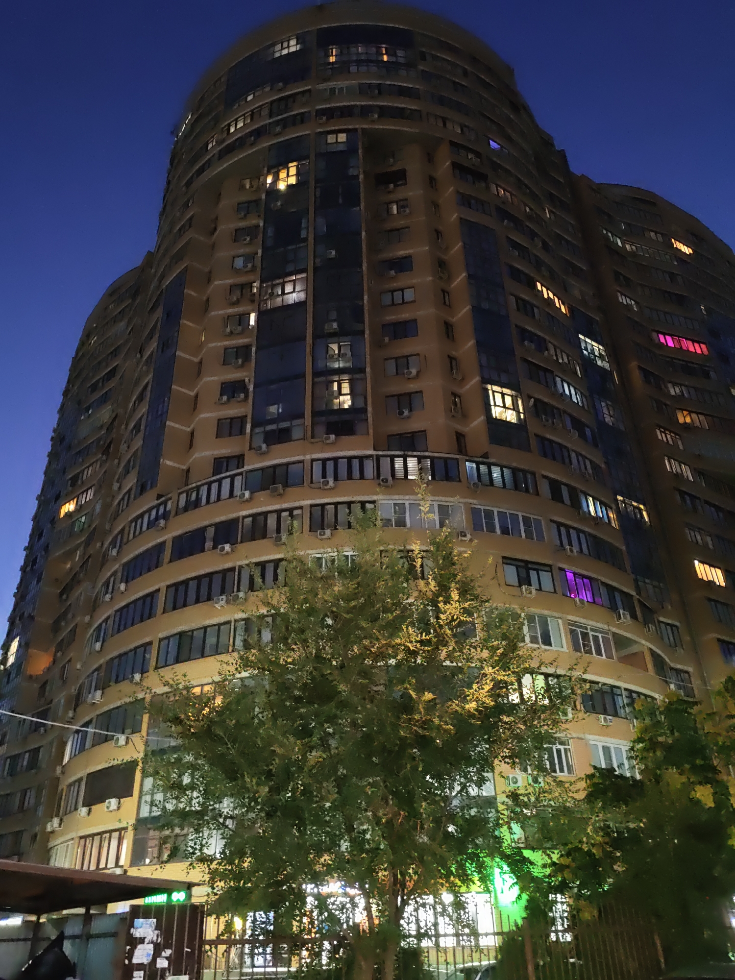 An upward-angle, nighttime photograph captures the exterior of a tall, curved, multi-story residential building in Rostov-on-Don, Russia. The building's facade is predominantly a pale yellow color, with dark blue glass panels and numerous windows, many of which are illuminated with warm yellow or orange light, indicating occupancy. Several air conditioning units are visible mounted on the exterior walls. In the foreground, the dense green foliage of trees partially obscures the lower floors of the building. Some of the trees appear illuminated by a bright green light emanating from a sign on the building's ground level. The sky above is a deep, clear blue, transitioning to darker shades of navy as it ascends, suggesting twilight or early night. The overall scene depicts a modern urban residential environment at dusk.