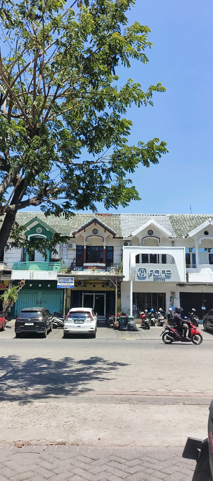 The image depicts a street scene in Kureksari, Indonesia, under a clear blue sky. In the foreground, a paved road with a brick border is visible, with shadows of a large tree cast across it. To the left, a substantial tree with green foliage partially obscures the view of the buildings.
The main focus is a row of commercial buildings. The first building on the left has a green facade and a white architectural detail at the top. Adjacent to it is a building with a cream-colored facade and a maroon-trimmed roof gable, featuring a circular window. Further to the right is a modern building with a white facade and a curved architectural element above the entrance, displaying the "Fore Coffee" logo in dark lettering. The buildings have tiled roofs.
Parked on the street are two cars: a black SUV on the left and a white SUV on the right. Several motorcycles are parked in front of the buildings, and one person is riding a red motorcycle in the middle of the street, moving towards the right of the frame. Trash bins and bags are visible near the motorcycles. The overall environment suggests a commercial area with pedestrian and vehicle traffic.
