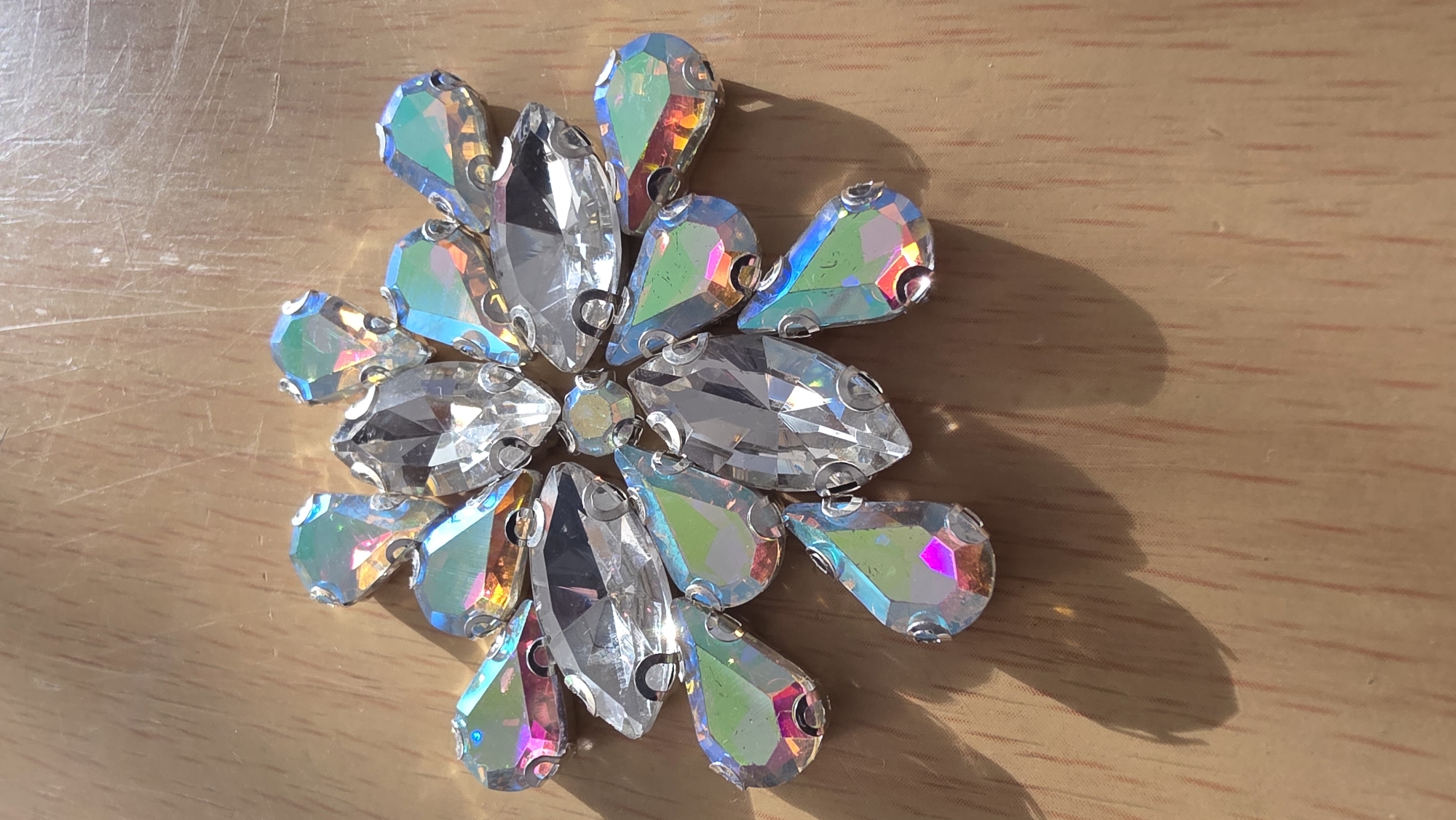 A close-up photograph shows an arrangement of shimmering jewels on a light brown, wood-grain surface. The jewels are teardrop-shaped, with a mixture of clear and iridescent colors. They are arranged in a radial pattern, resembling a flower or a starburst. The lighting suggests the objects are in direct sunlight. There is no indication of this image being taken in Blagoevgrad, Bulgaria or any other specific location. There are no people present in the image.