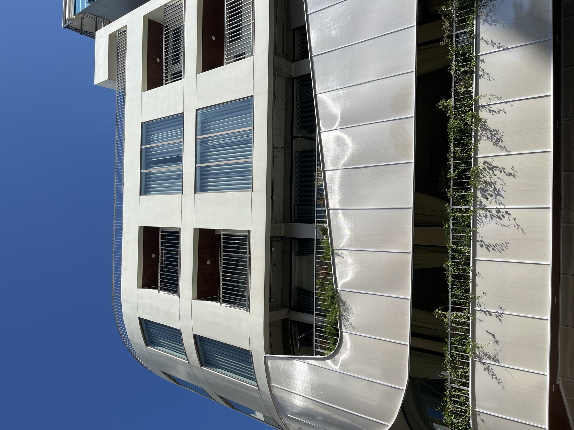 A low-angle, off-level shot shows a tall, modern building against a clear blue sky. The building has two distinct sections. The left section is white with square windows and horizontal metal blinds. The windows are stacked vertically. Between the rows of windows are open balconies with metal bars. The section bends forward in a gentle curve. The right section is covered with vertical, translucent panels that curve around a set of plants growing on a metal trellis. The building’s location in the city of Đà Nẵng, Vietnam, cannot be determined from the image. There are no people visible in the image.