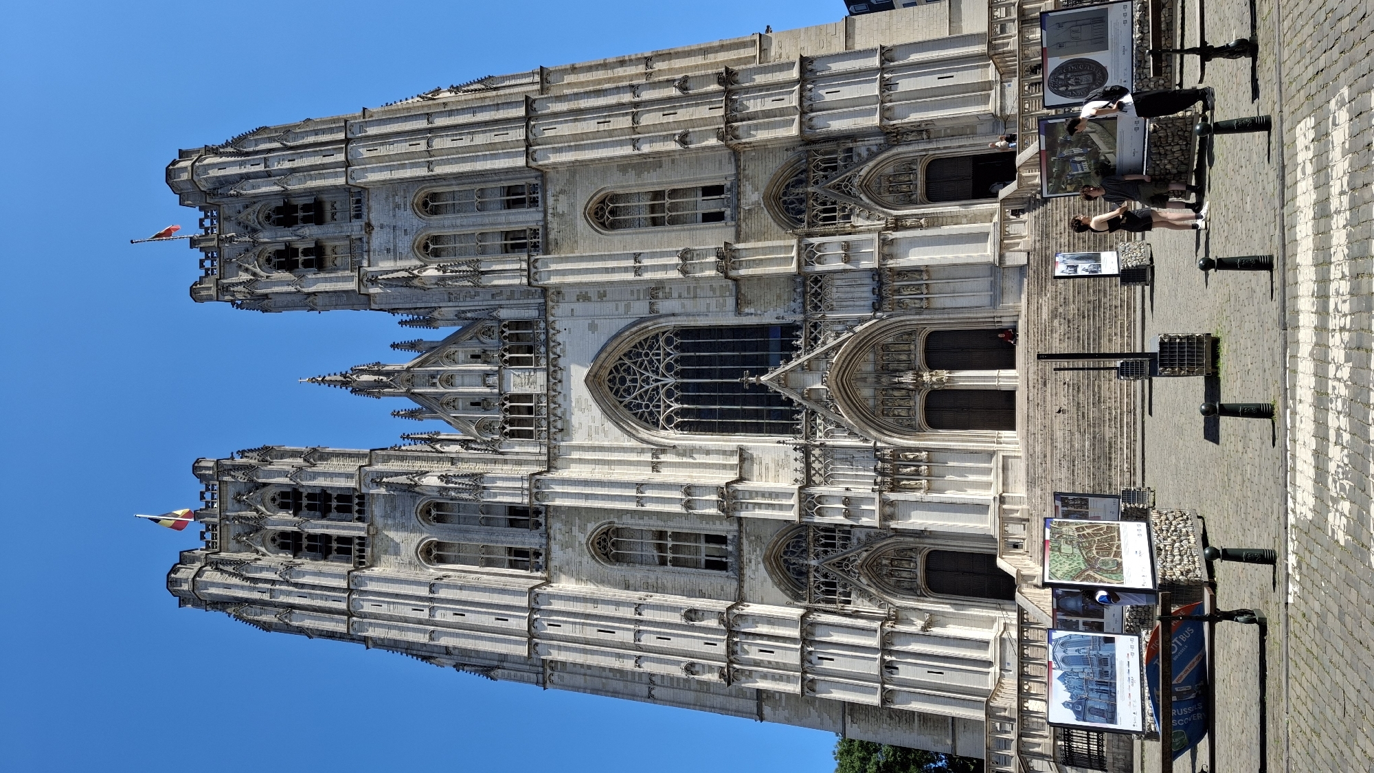 This is a full-body, low-angle shot of the exterior of a large, ornate Gothic cathedral. The cathedral is made of light-colored stone and features two prominent bell towers with intricate carvings and arched windows. A Belgian flag flies from the top of the left tower, and another flag flies from the right tower. The facade of the cathedral has a large, arched stained-glass window above a set of double doors, with numerous sculptures and decorative elements surrounding the entrance.
In the foreground, a wide set of stone stairs leads up to the cathedral entrance. Scattered around the base of the stairs and the paved plaza in front of them are several large display boards showcasing images of the cathedral and local maps. A few people are visible in the scene: three young adults are standing and talking near the display boards on the right, and a single person is seated on the steps of the cathedral. The plaza is paved with cobblestones, and black bollards line the pathway. The sky is a clear, vibrant blue.